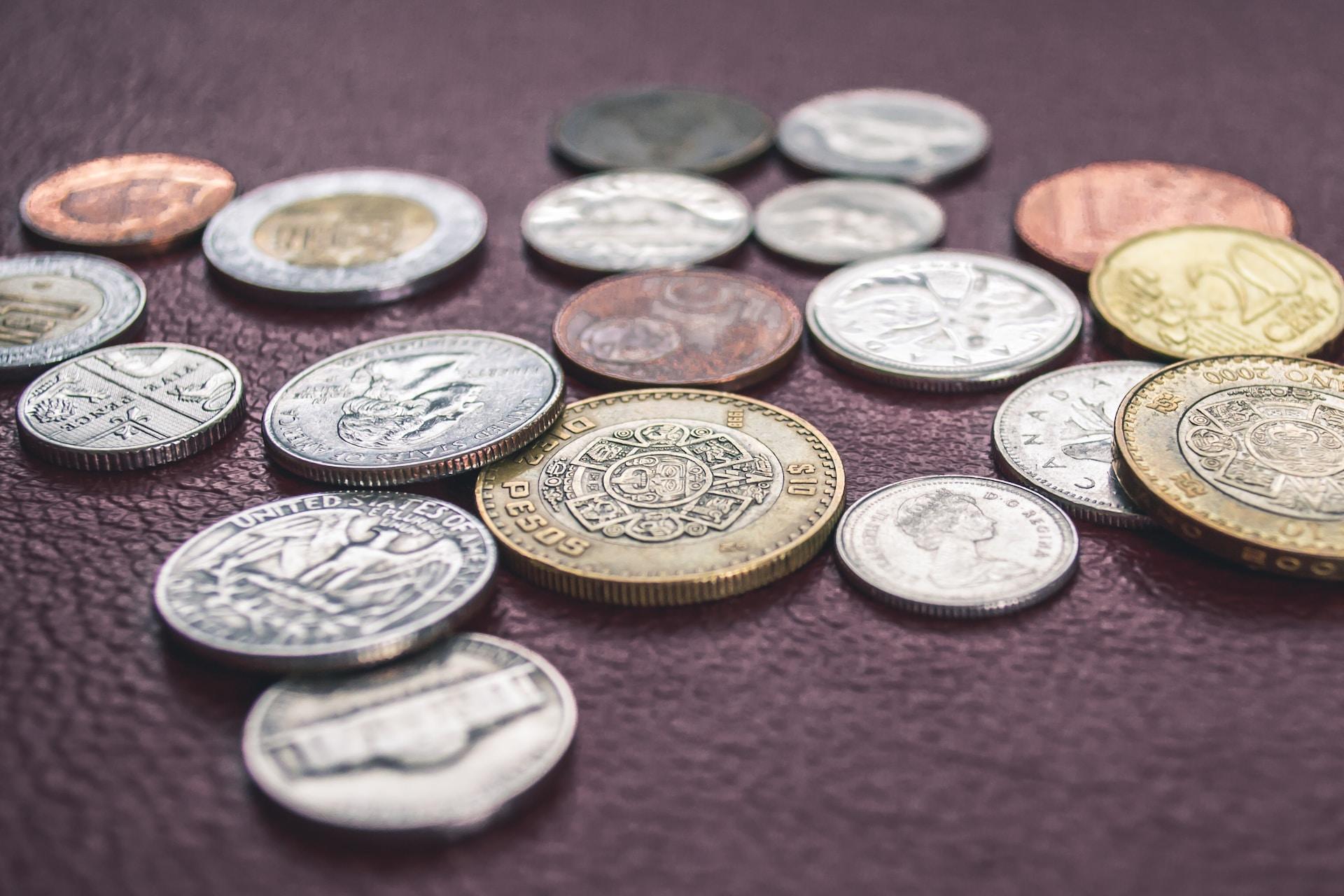 A scattering of different currency coins from various countries, laying on a reddish-brown surface.