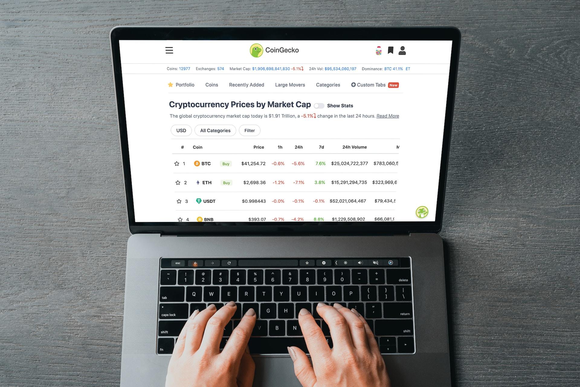 A silver laptop with a black keyboard sits on a greyish wooden surface. It is open, it's screen displaying an array of cryptocurrency prices. 
