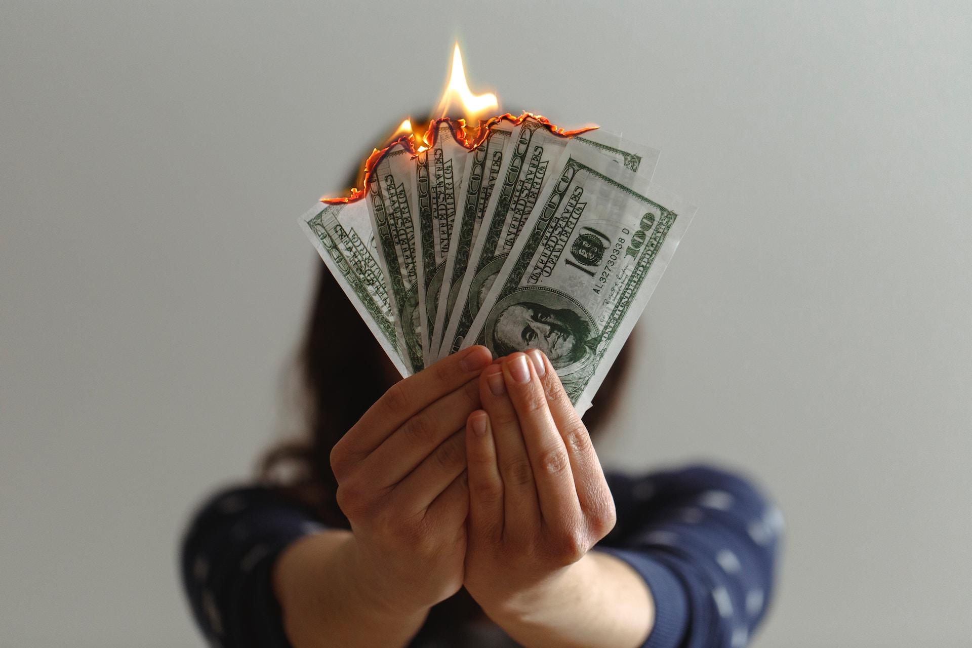 A person wearing a dark blue top with the sleeves rolled up holds a fan of $100 bills whose edges are in flames. 