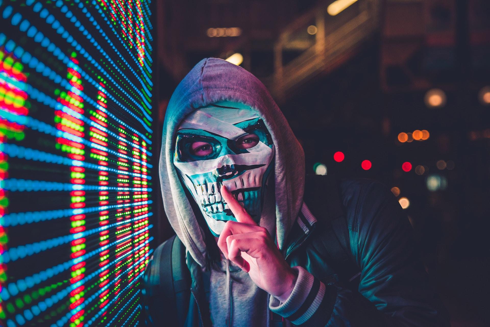 A person in a multicoloured skull mask and a grey hoodie covering their head places their finger on their mouth in the shushing gesture as they stand next to a green, blue and red plxelated light display at night. 