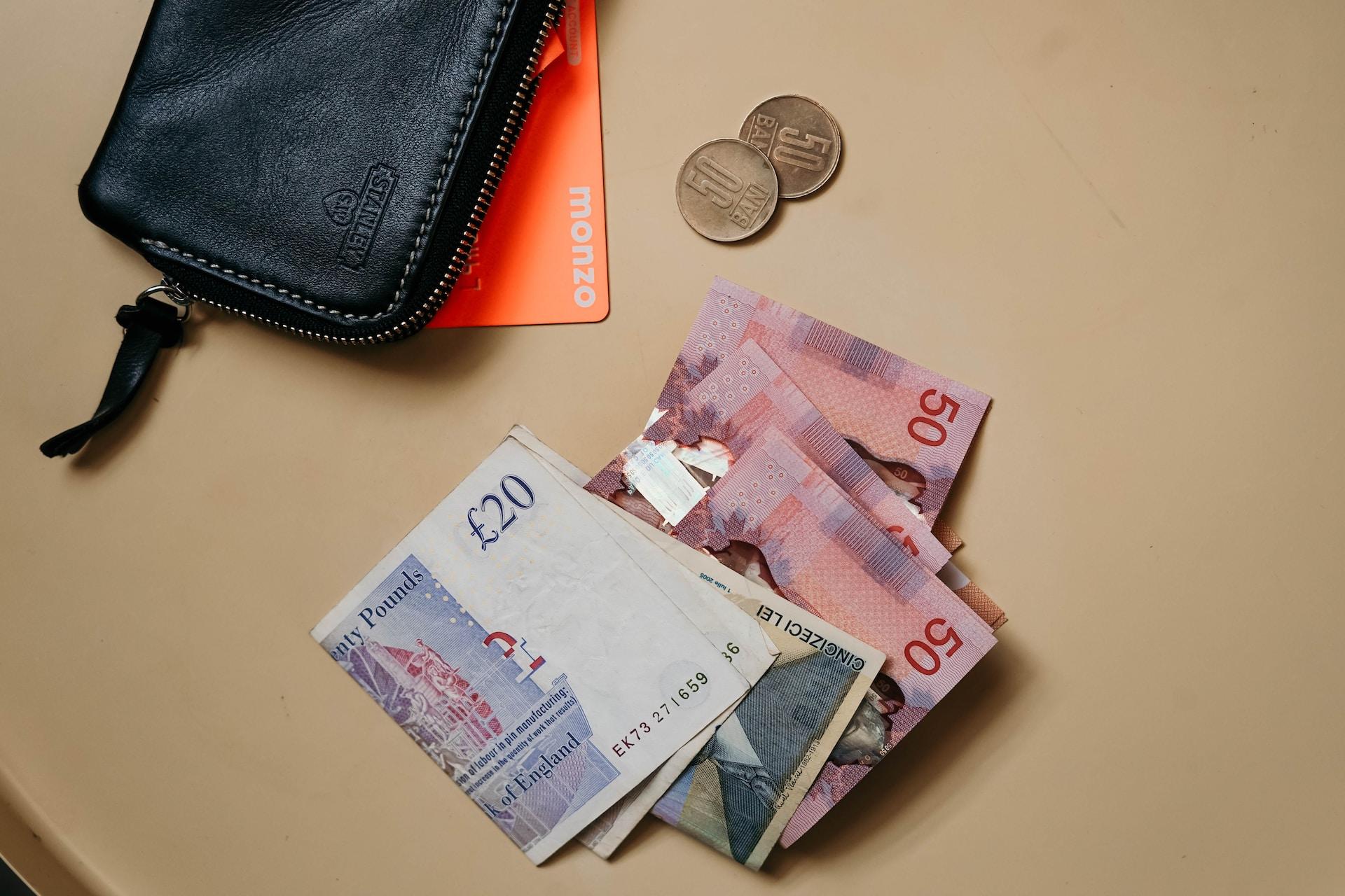 A black wallet laying on a brown surface with an orange money card tucked under it and some bills and coins laying next to it.