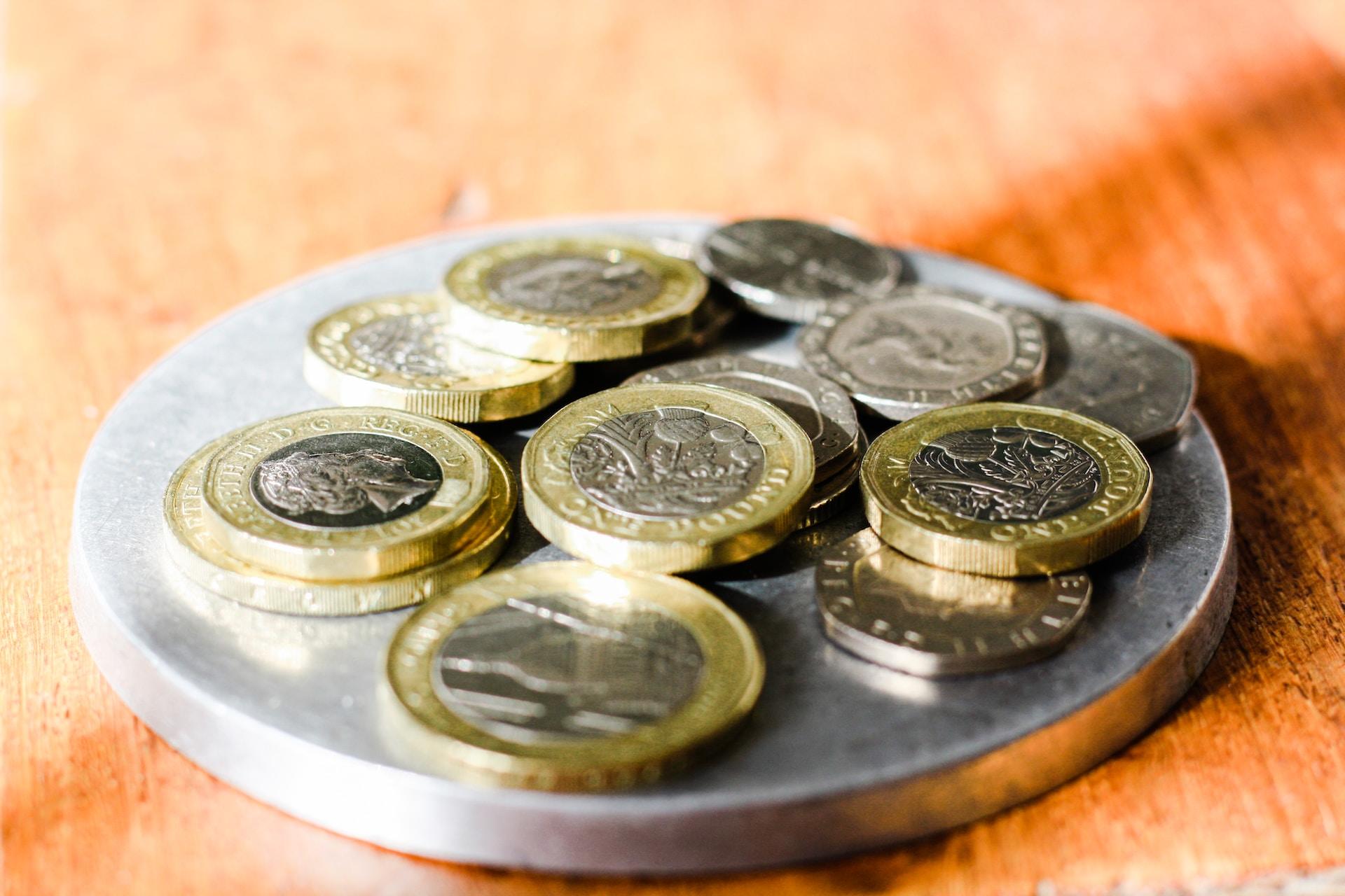 An assorted pile of GBP coins of various denominations placed atop a larger silver disc which, in turn, sits on a glossy wooden surface. 