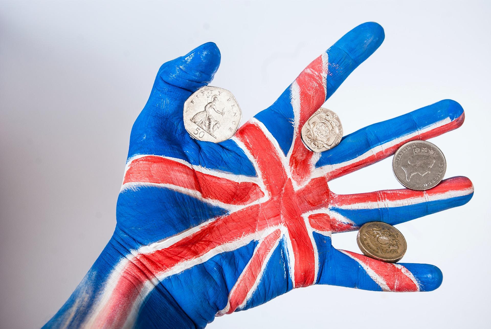 A human left hand with the thumb at the top and the palm facing the camera, painted like the British flag, holds a British coin between each of its fingers.