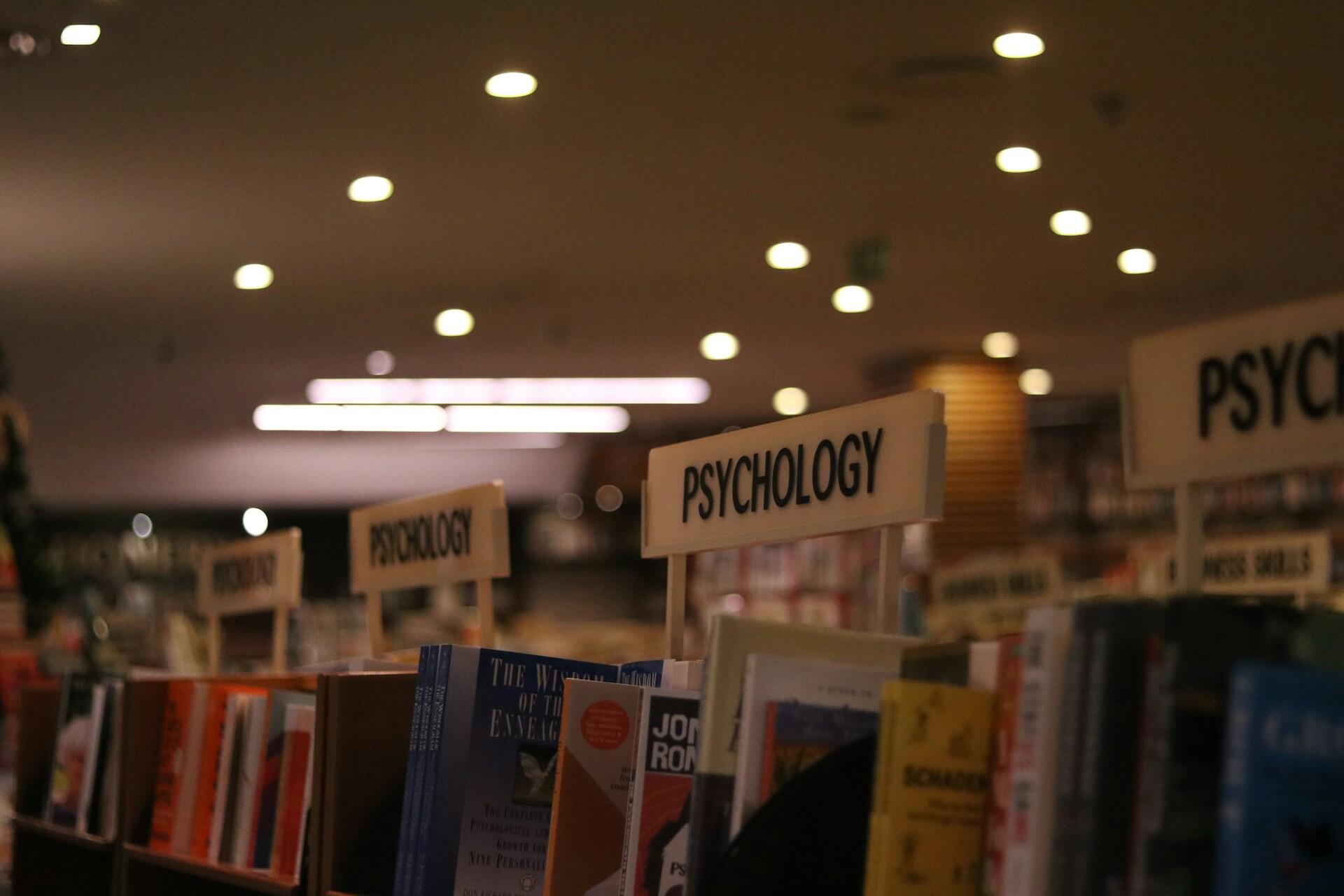 Bookshelves in a library or bookstore with multiple sections labeled “Psychology,” displaying psychology books under warm indoor lighting.
