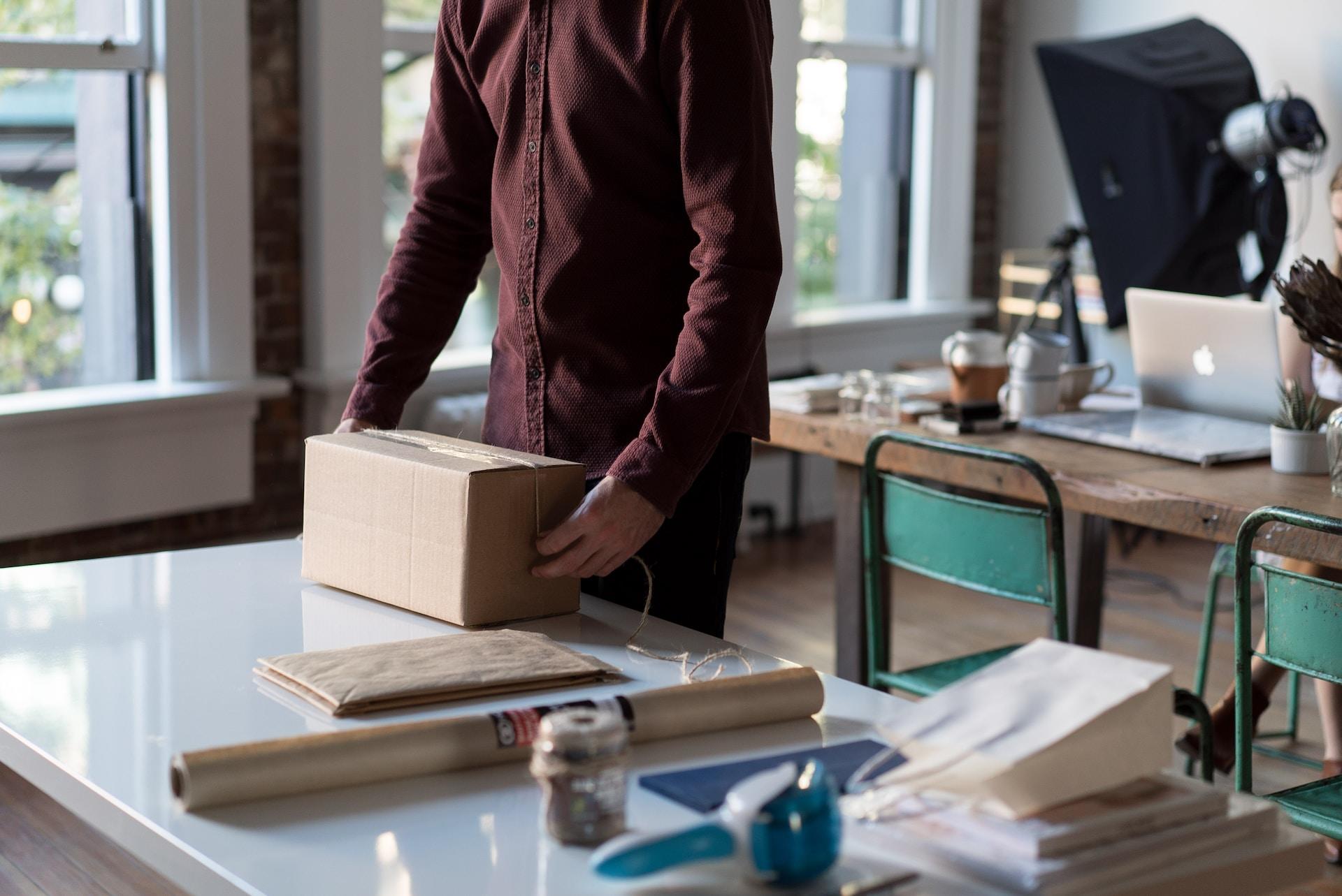 A person wearing a long sleeved brown shirt stands at a white table, tying a length of sisal rope around a package, with a roll of parcel paper and other office supplies next to him on the table. 