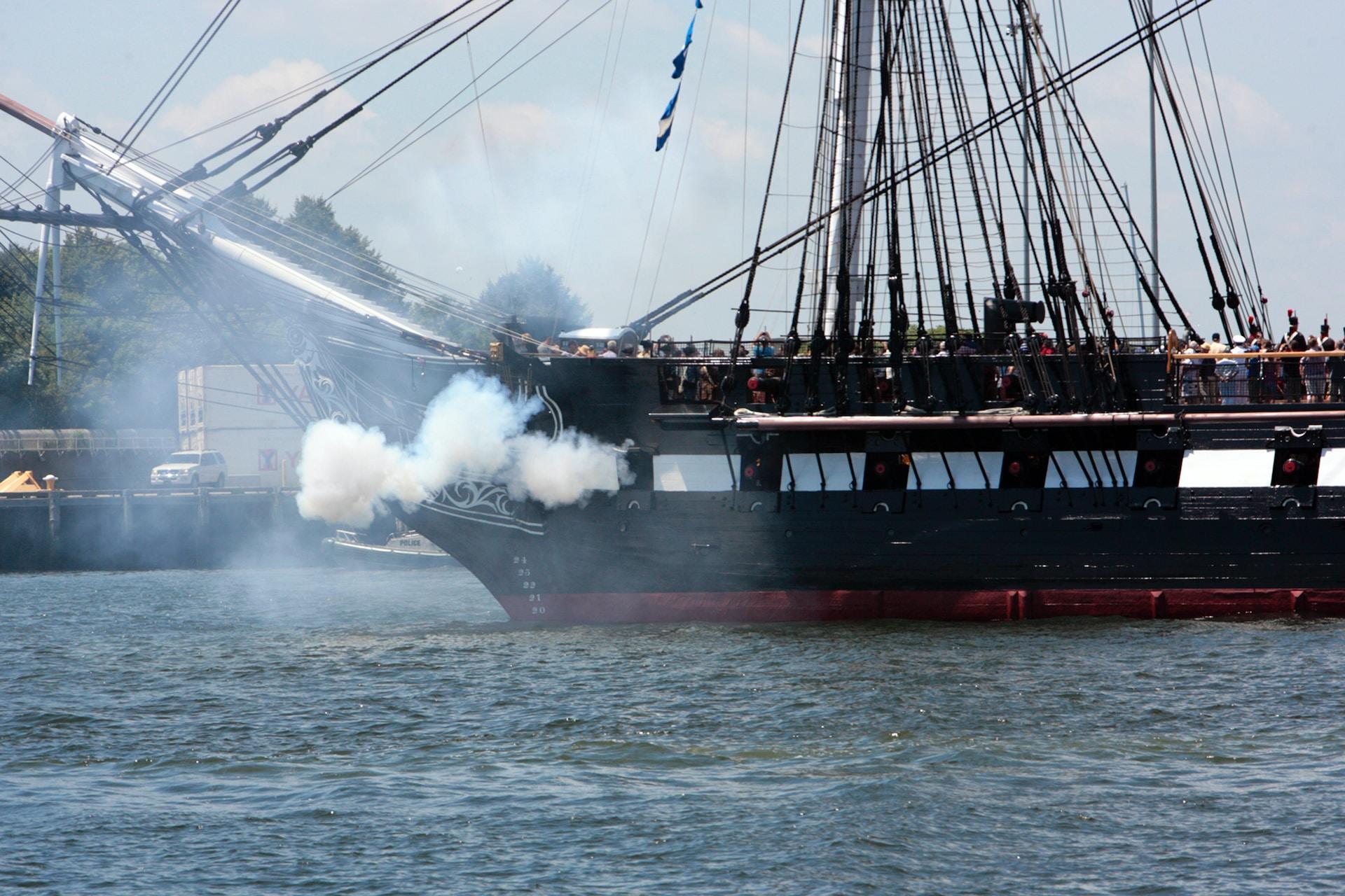 A black galleon ship with a red keel has its sails down as it fires a shot across the water. 