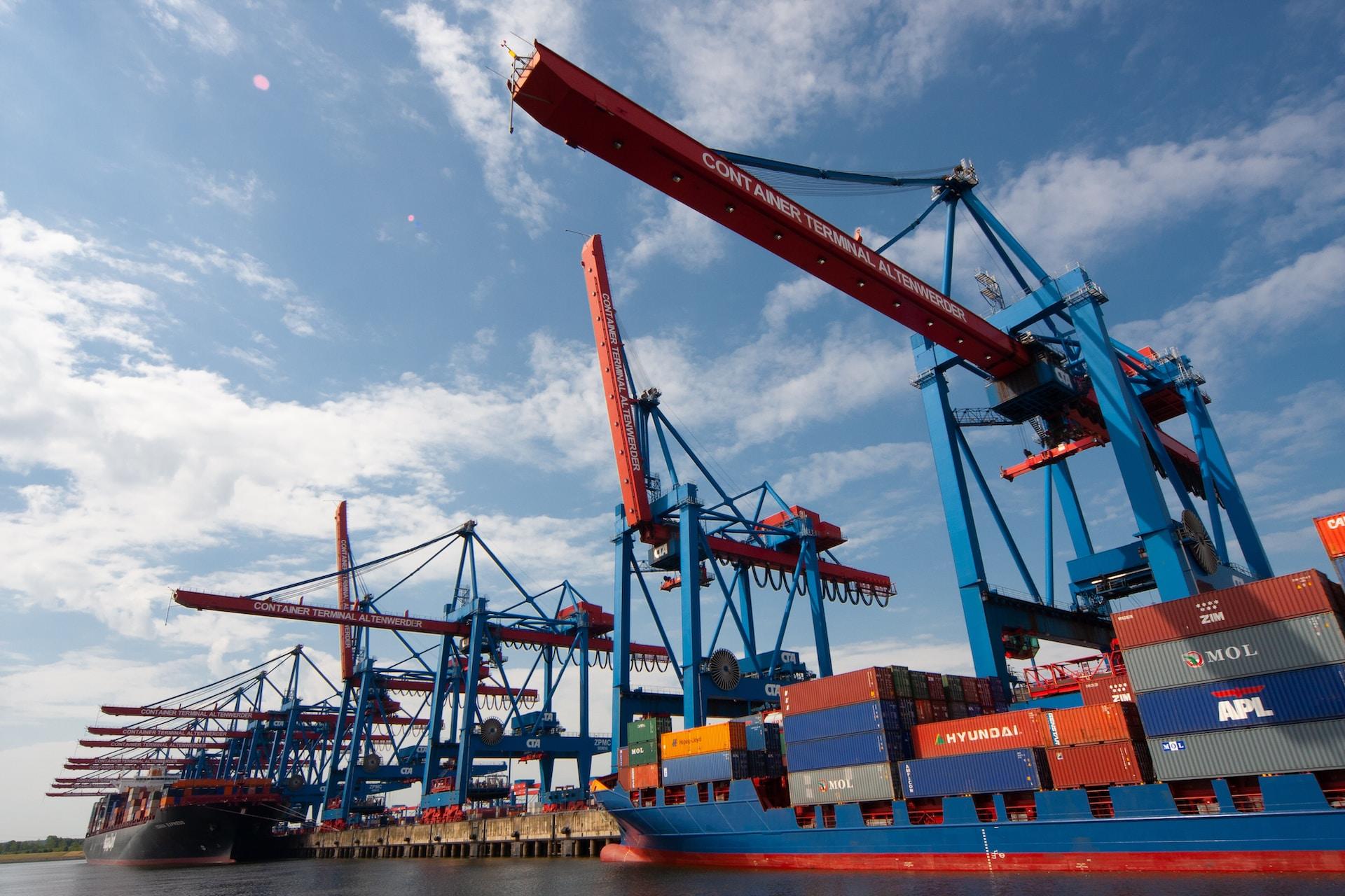 A shipping port with two container ships docked; one blue and the other black. Along the dock is a line of blue and orange cranes. 