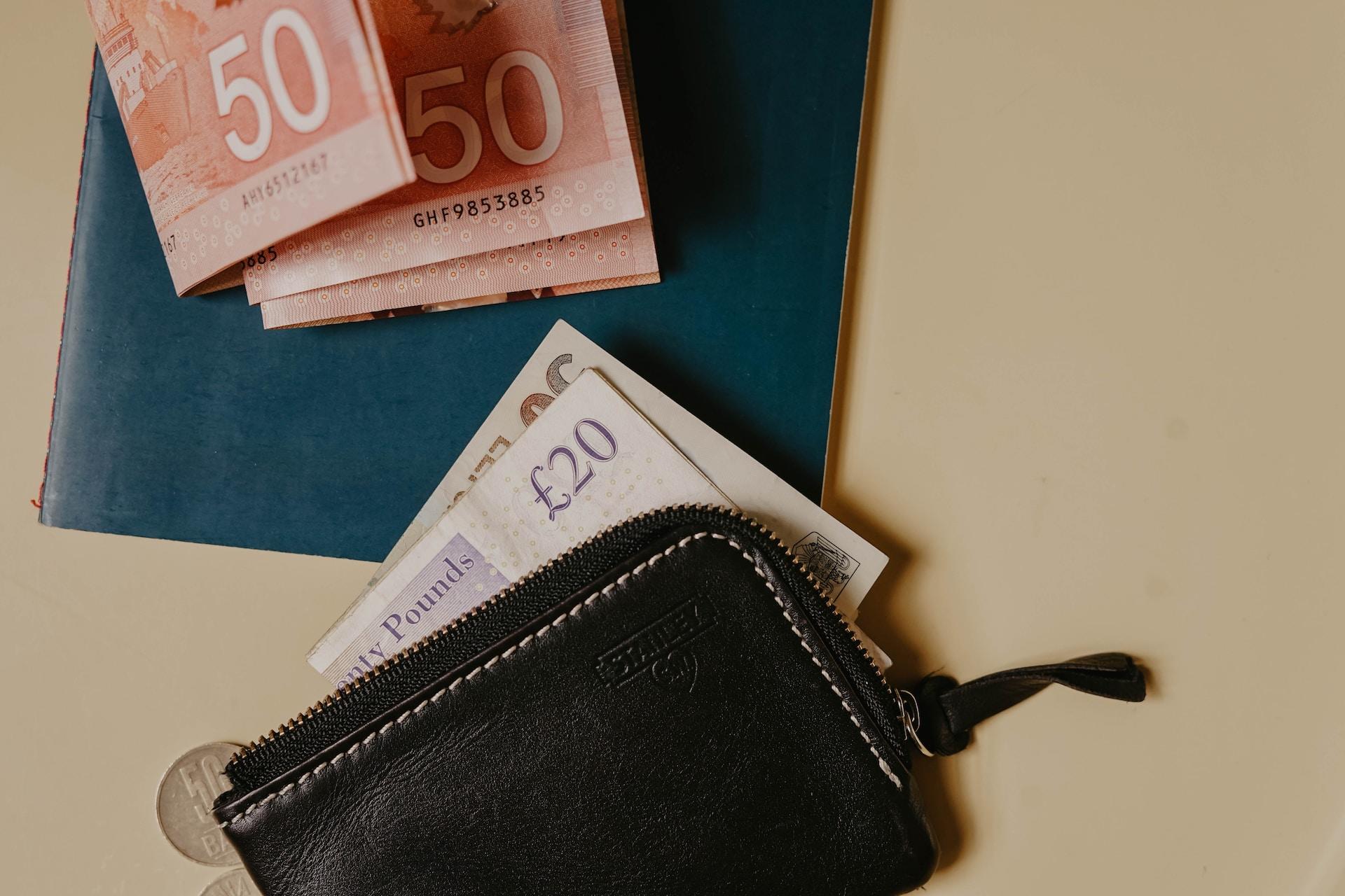 A black wallet lays unzipped on a light-coloured surfaced next to a small turquoise mat, upon which lay several folded £50 bills, while a £20 bill protrudes from the wallet.