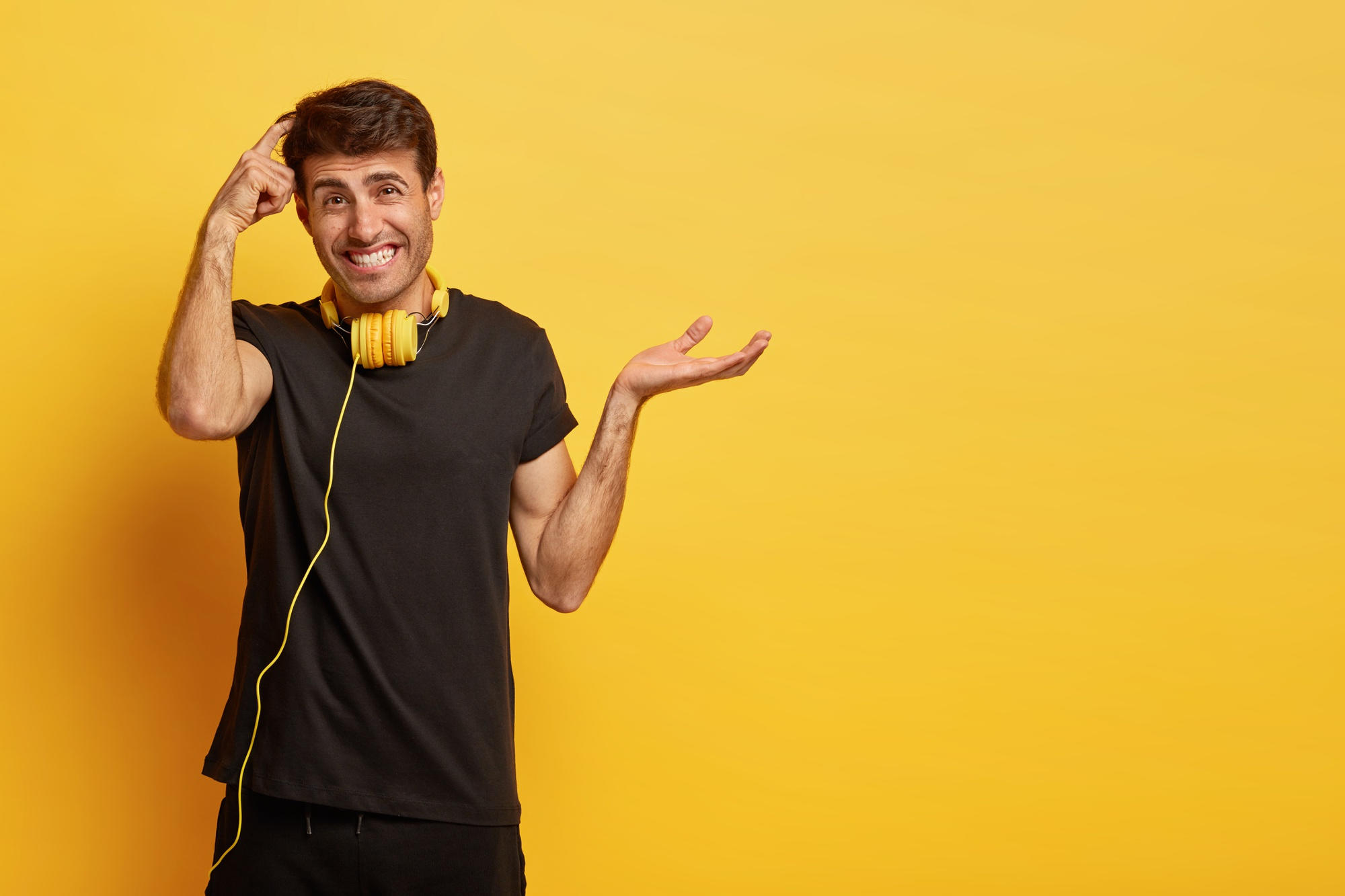 A man wearing black clothes with yellow headphones around his neck is very confused and stands in front of a yellow wall