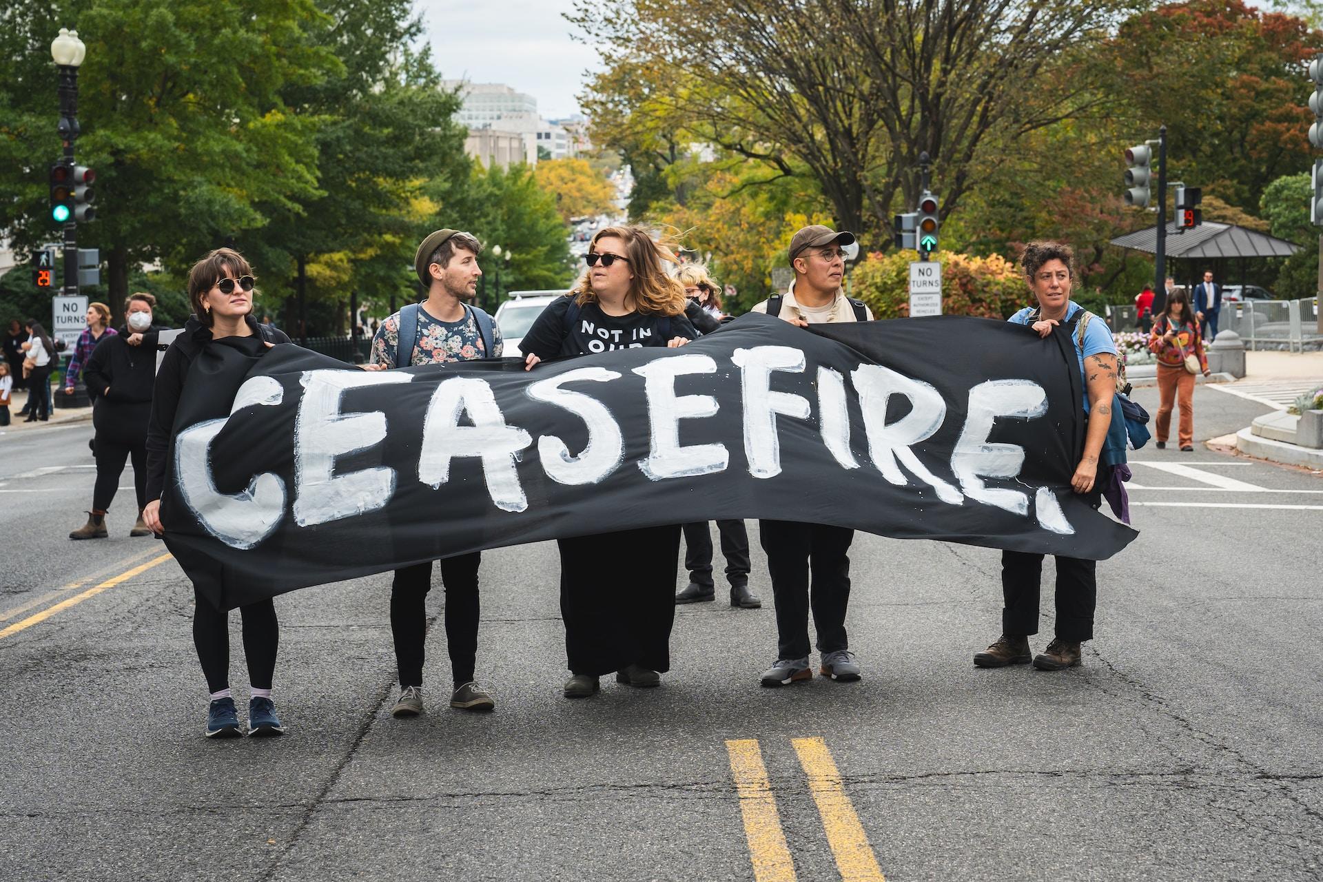 Five people dressed in casual clothing hold a large black banner with 'ceasefire' painted on it in white.
