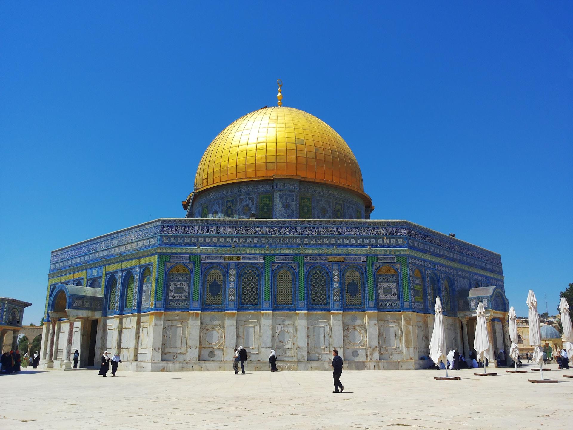 The Al-Aqsa Mosque in the Old City of Jerusalem, Palestine with a golden dome, and blue and white tiled walls and a few faithful walking about in front of it on a sunny day.