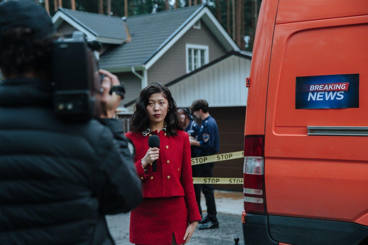 A reporter stands in front of a scene holding a microphone and looking into the camera. A news van is parked next to her.
