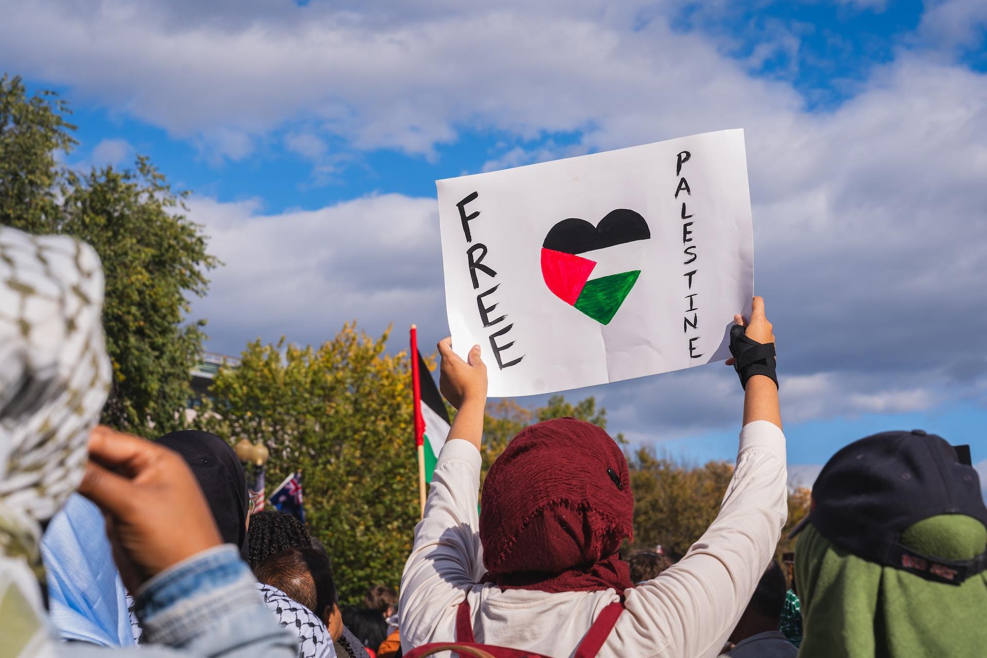 A person wearing a red headscarf and a white top, carrying a red backpack, holds up a sign that says 'free Palestine' with the Palestinian flag in the shape of a heart, in a demonstration on a sunny day.