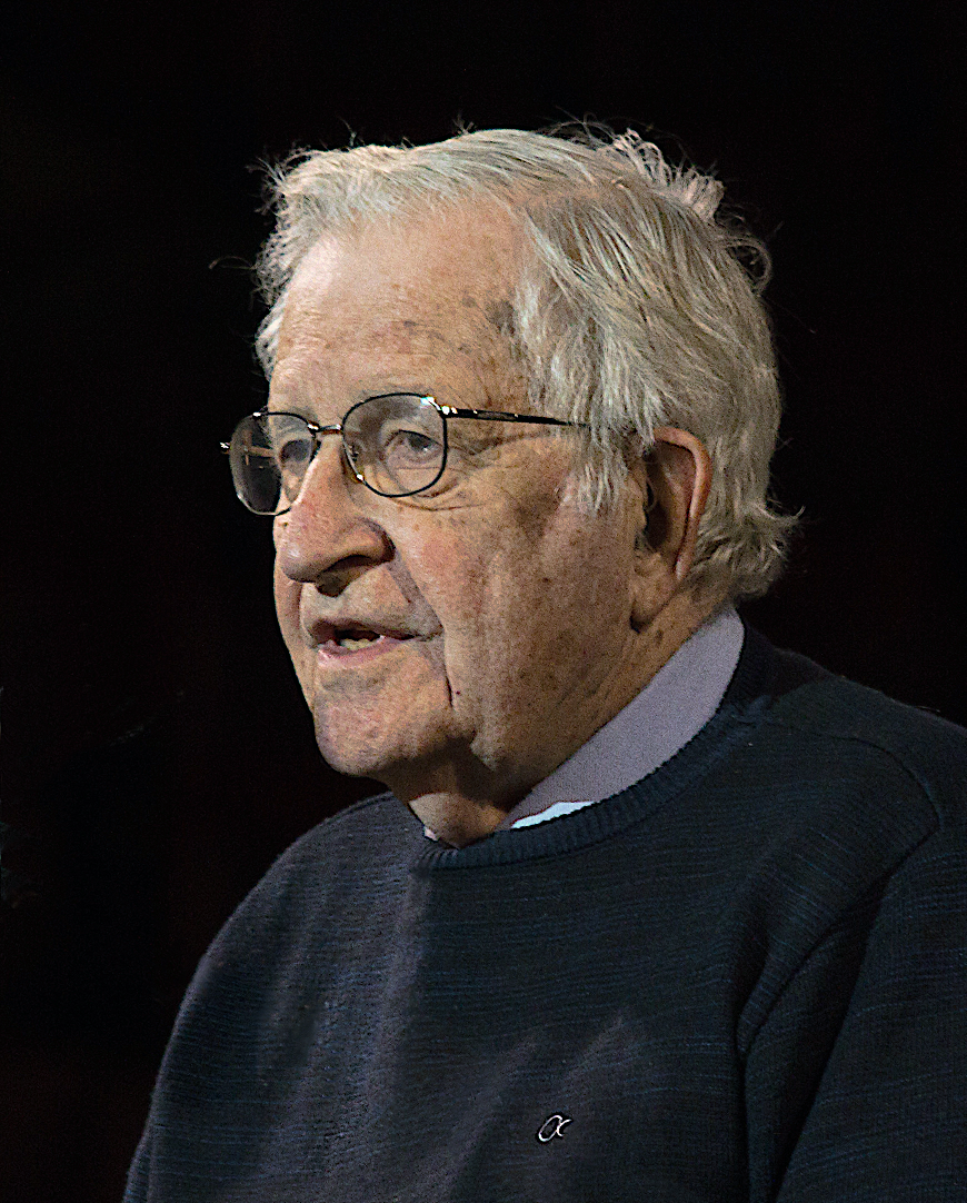 A portrait of Noam Chomsky wearing a grey jumper over a white collared shirt, and silver glasses against a black background. 