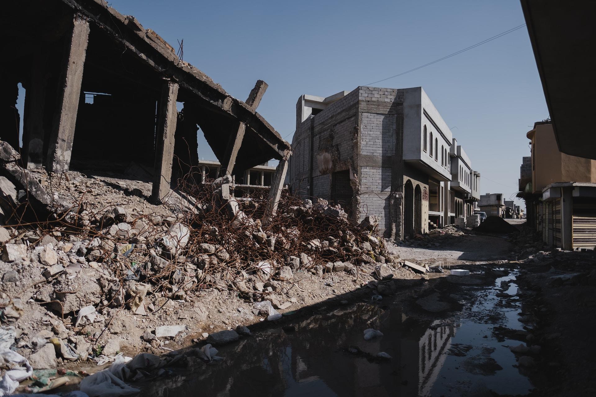 A bombed-out structure with pillars askew poking out of the rubble to support what's left of a roof. The street fronting it is deserted and a large puddle reflects the damage.