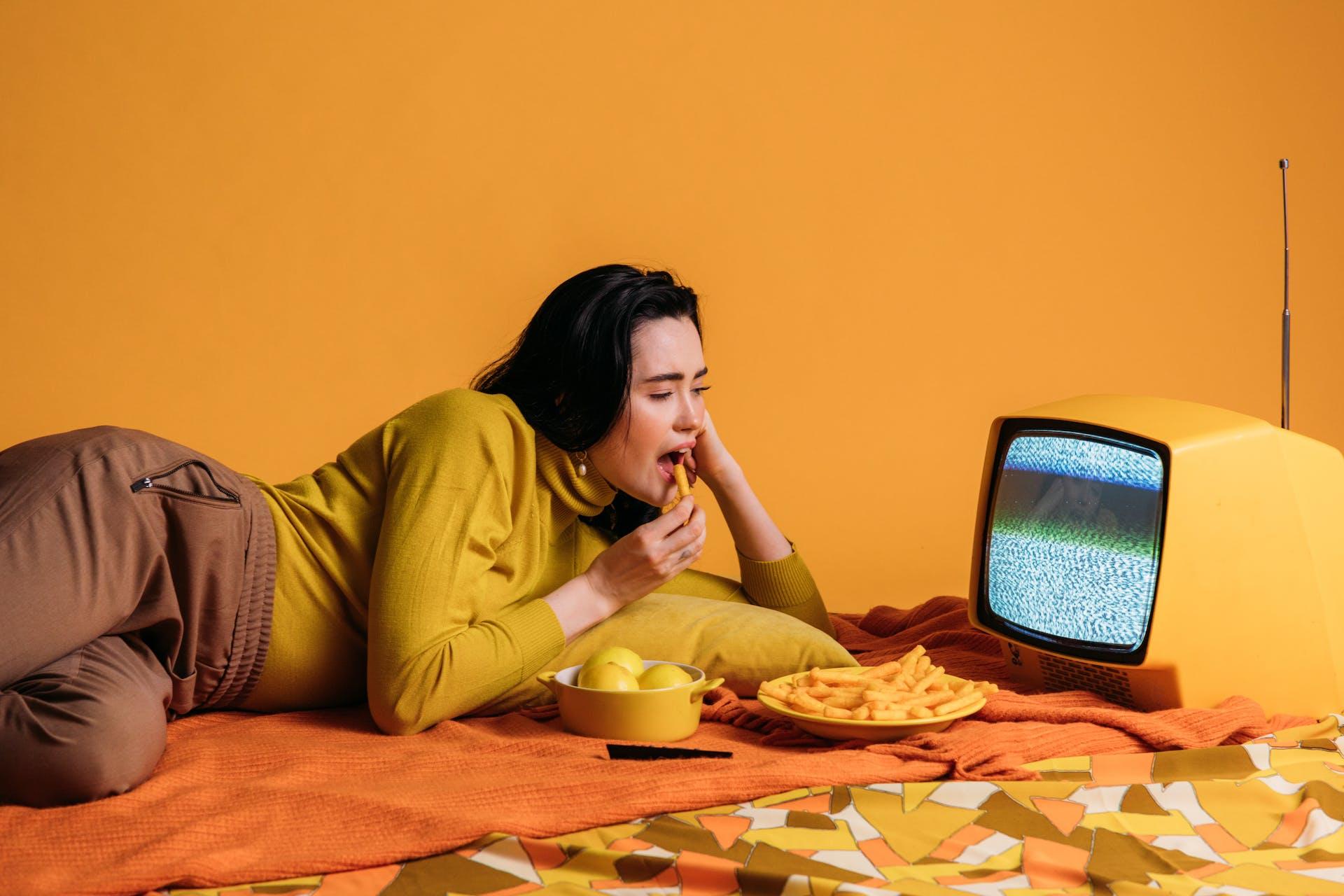 A girl in a yellow sweater eating cheesy snacks on an orange blanket in a yellow room