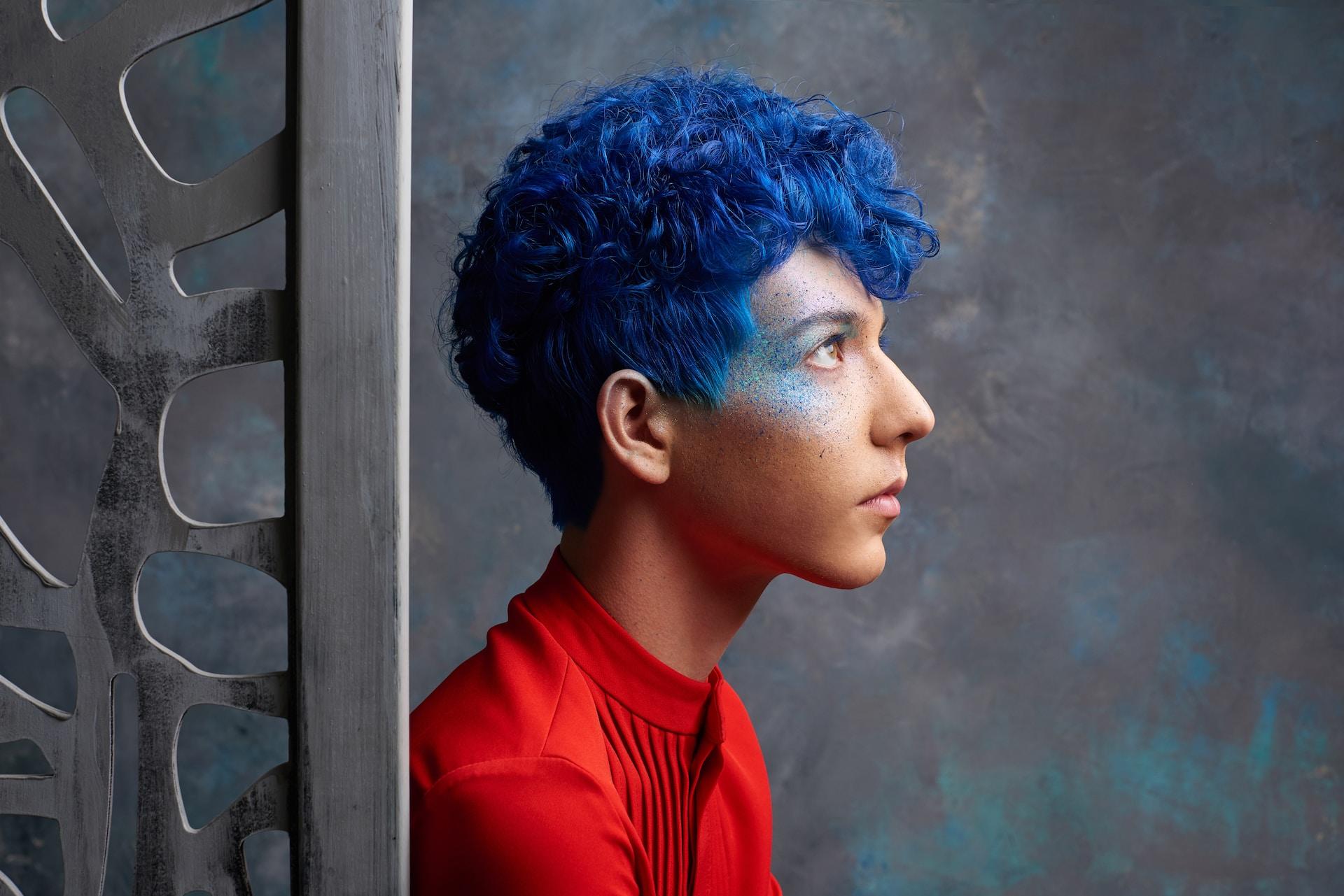 A youth with royal blue hair, wearing a red shit, stands in profile against a blue and grey background. 