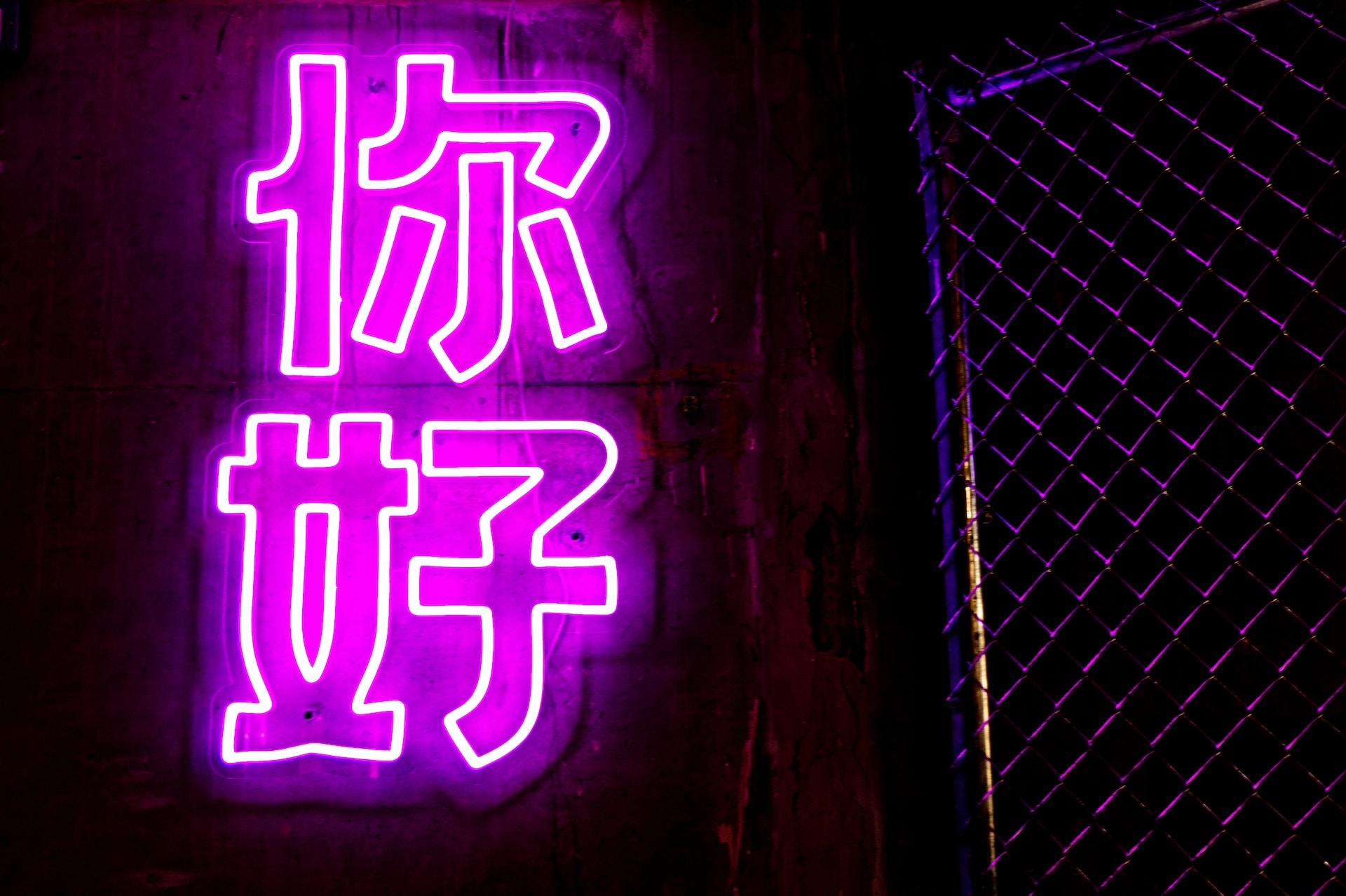 A purple neon sign displaying the Chinese characters for 'hello', hung next to a chain-link fence.  