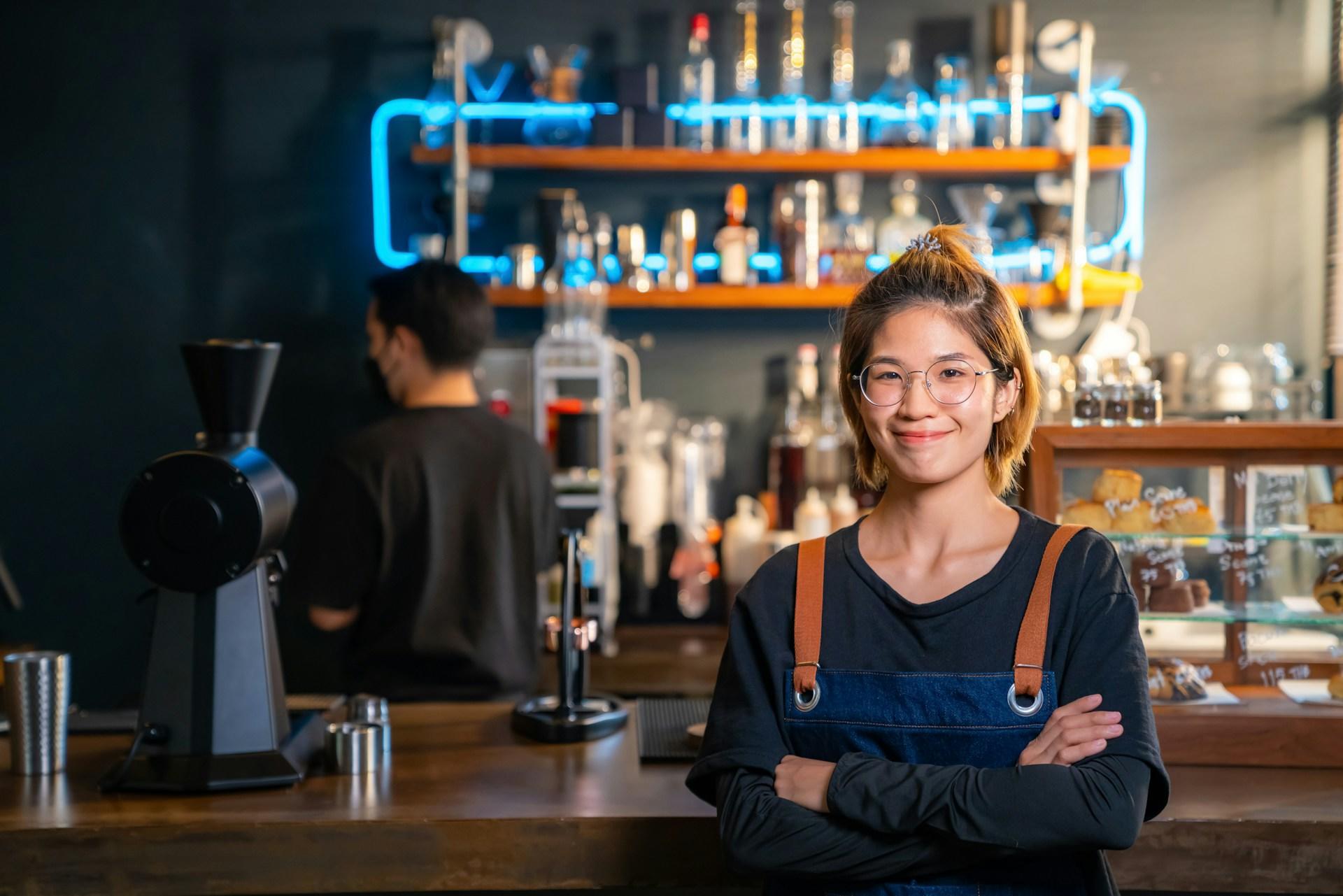 A barista wearing wire-framed glasses and a denim apron over a dark shirt smiles at the camera as they stand in front of the bar. 