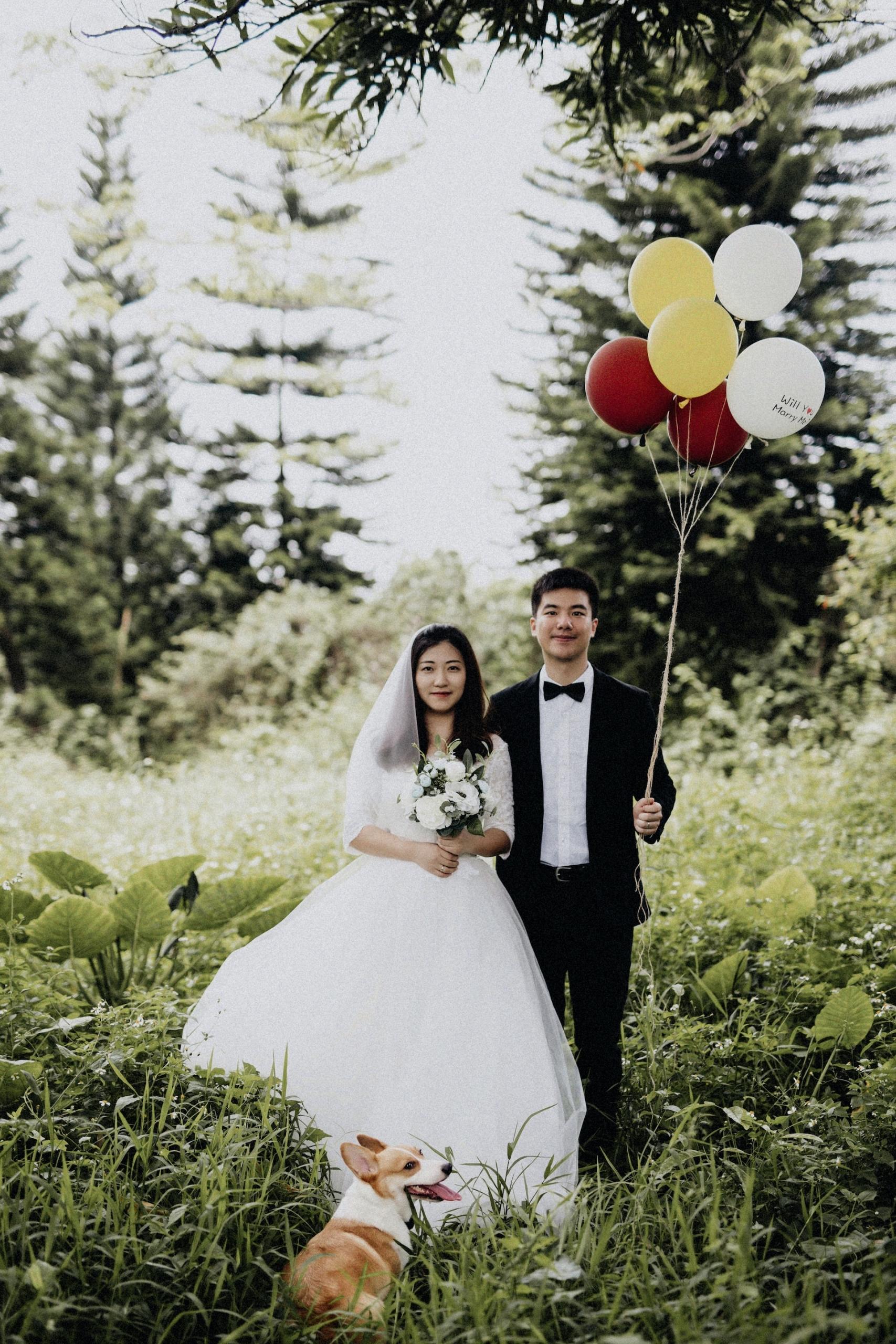 A man and woman wearing wedding garb stand side by side in a lush green field with slight smiles on their faces. The man holds the strings to a bunch of  balloons coloured red, yellow and white. A tan and white dog lays in front of the woman.  