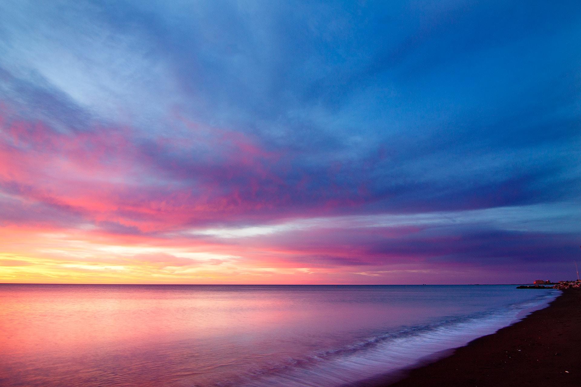A beach scene at sunset, with the sky's colors ranging from dark blue to yellow. 
