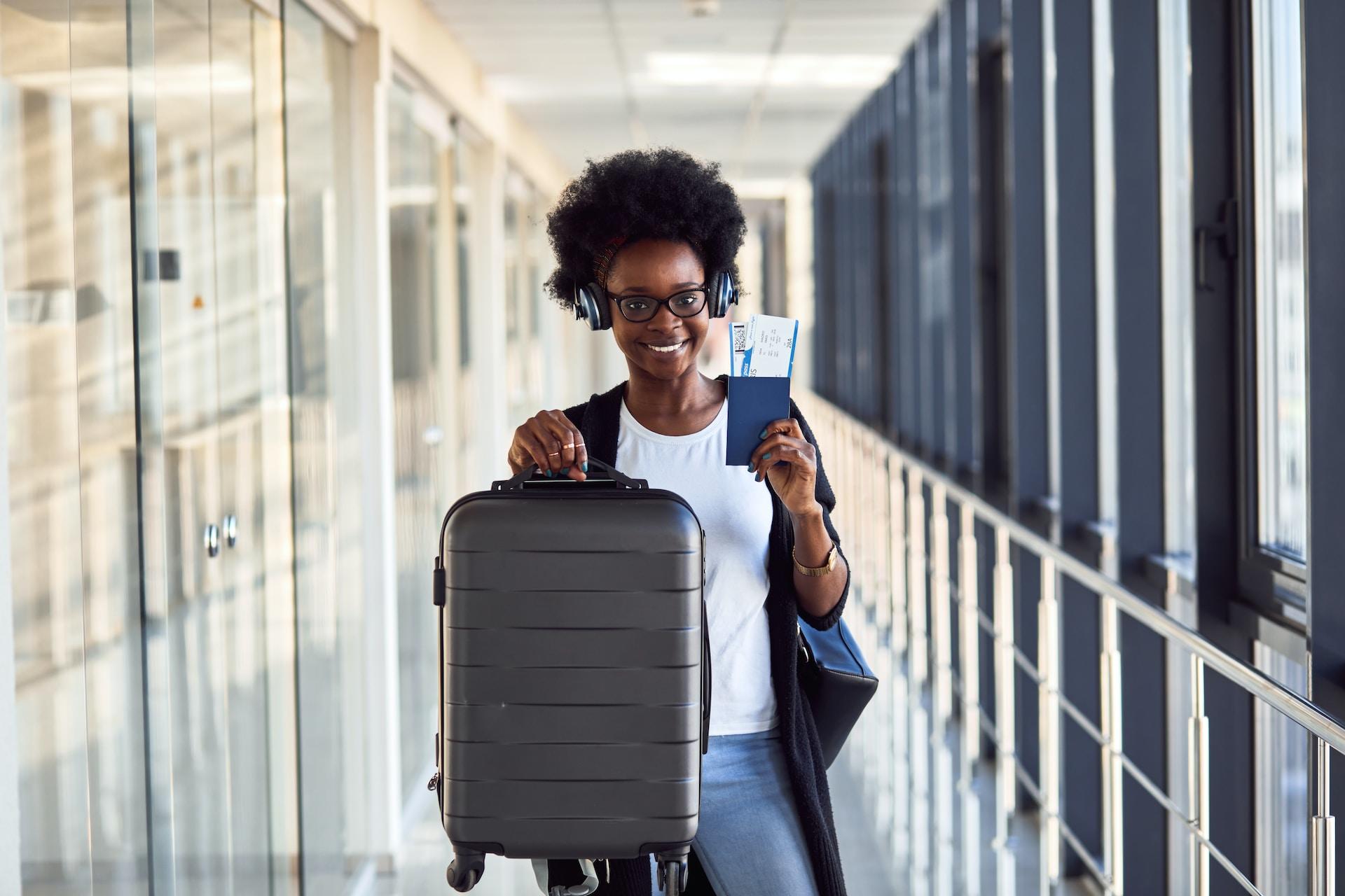A person wearing dark framed glasses and headphones smiles as they hold up their passport with airline tickets tucked in it and a grey suitcase.