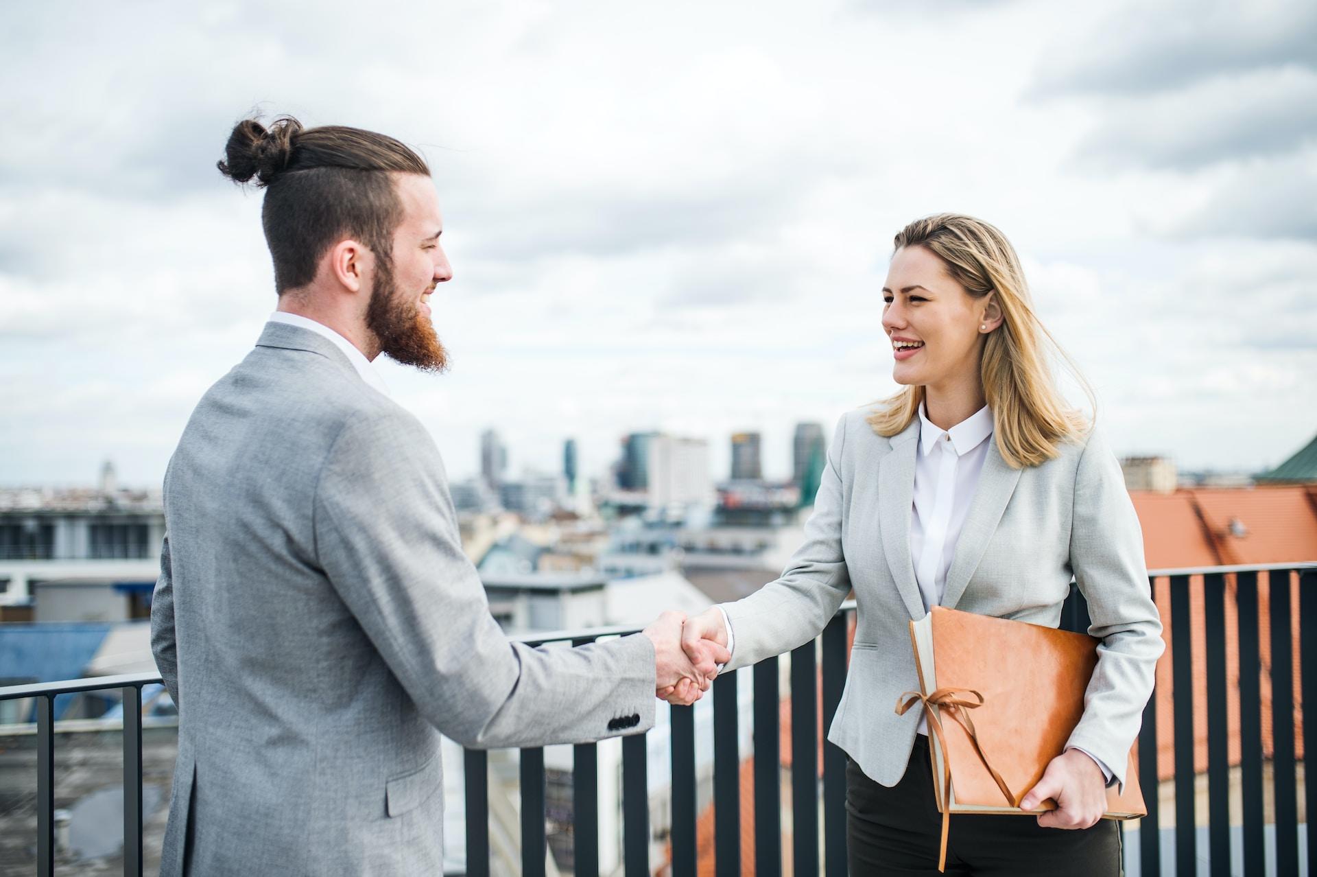 Two people wearing light grey business attire and white shirts meet on a bridge and shake hands. 