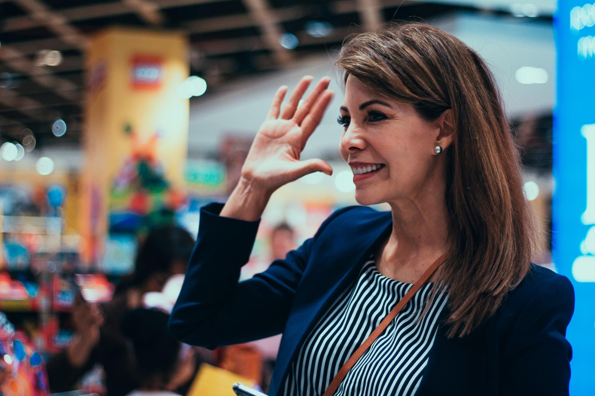 A person wearing a dark blazer over a green and white striped top smiles and holds up their right hand to wave hello.