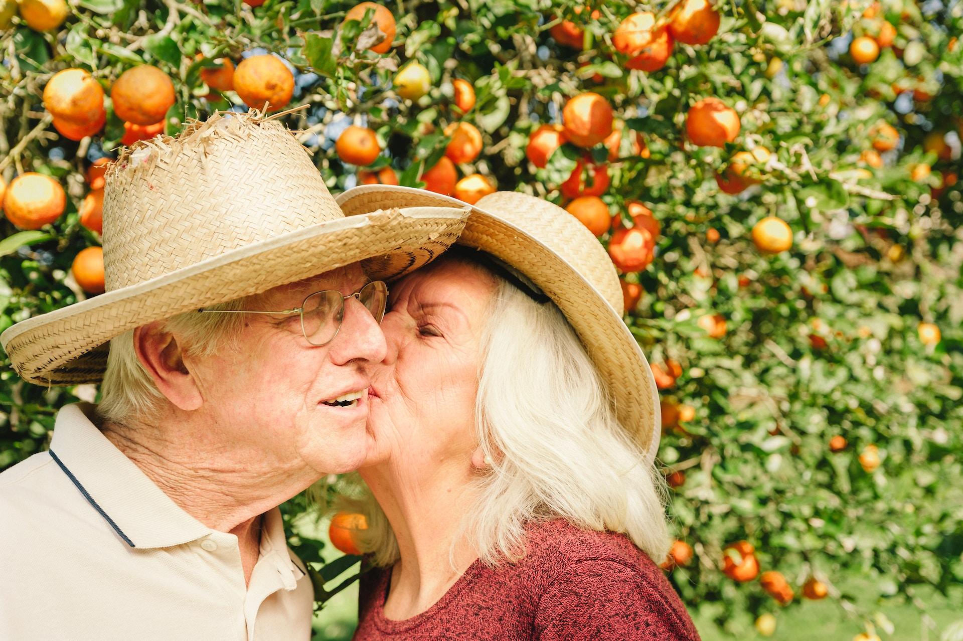A woman wearing a patterned reddish top and a straw hat kisses a man wearing a tan shirt and a straw hat on the cheek.