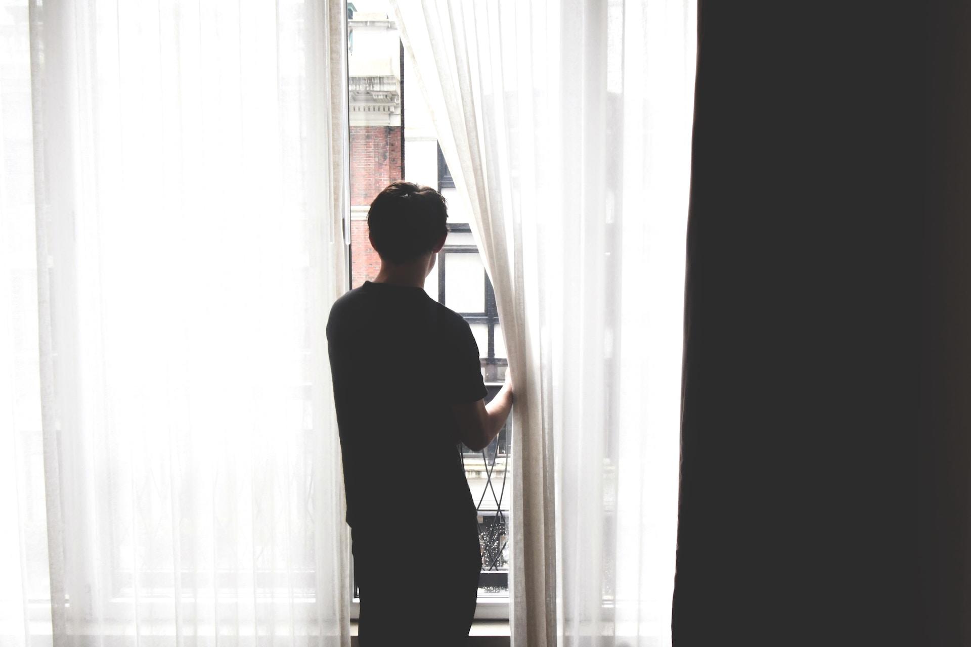 A person stands at a window which is covered by white sheer panels, one of which they have moved aside to look out the window.