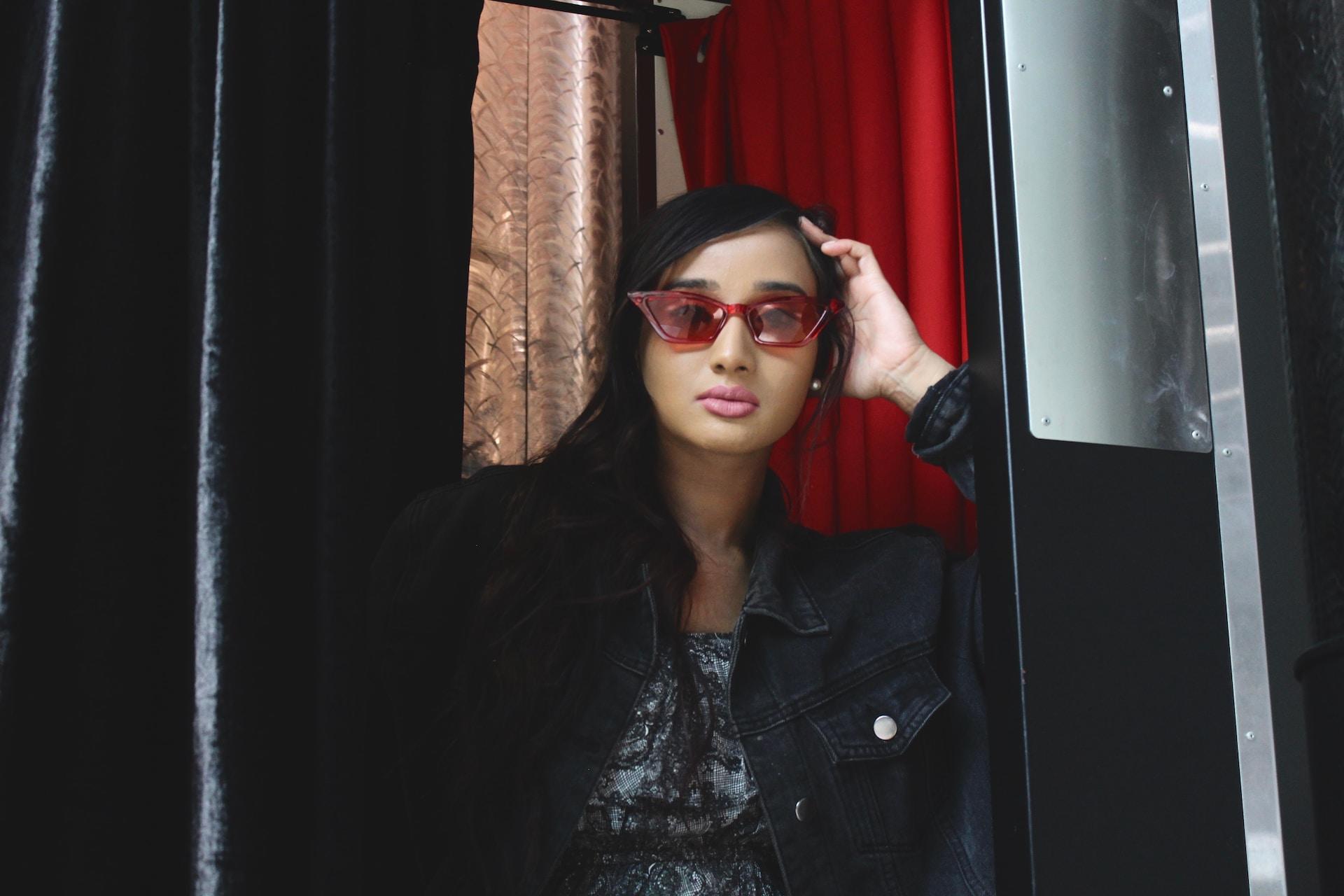A stylish young woman with long black hair wear dramatic rose-colored glasses as she leans against a black door. 