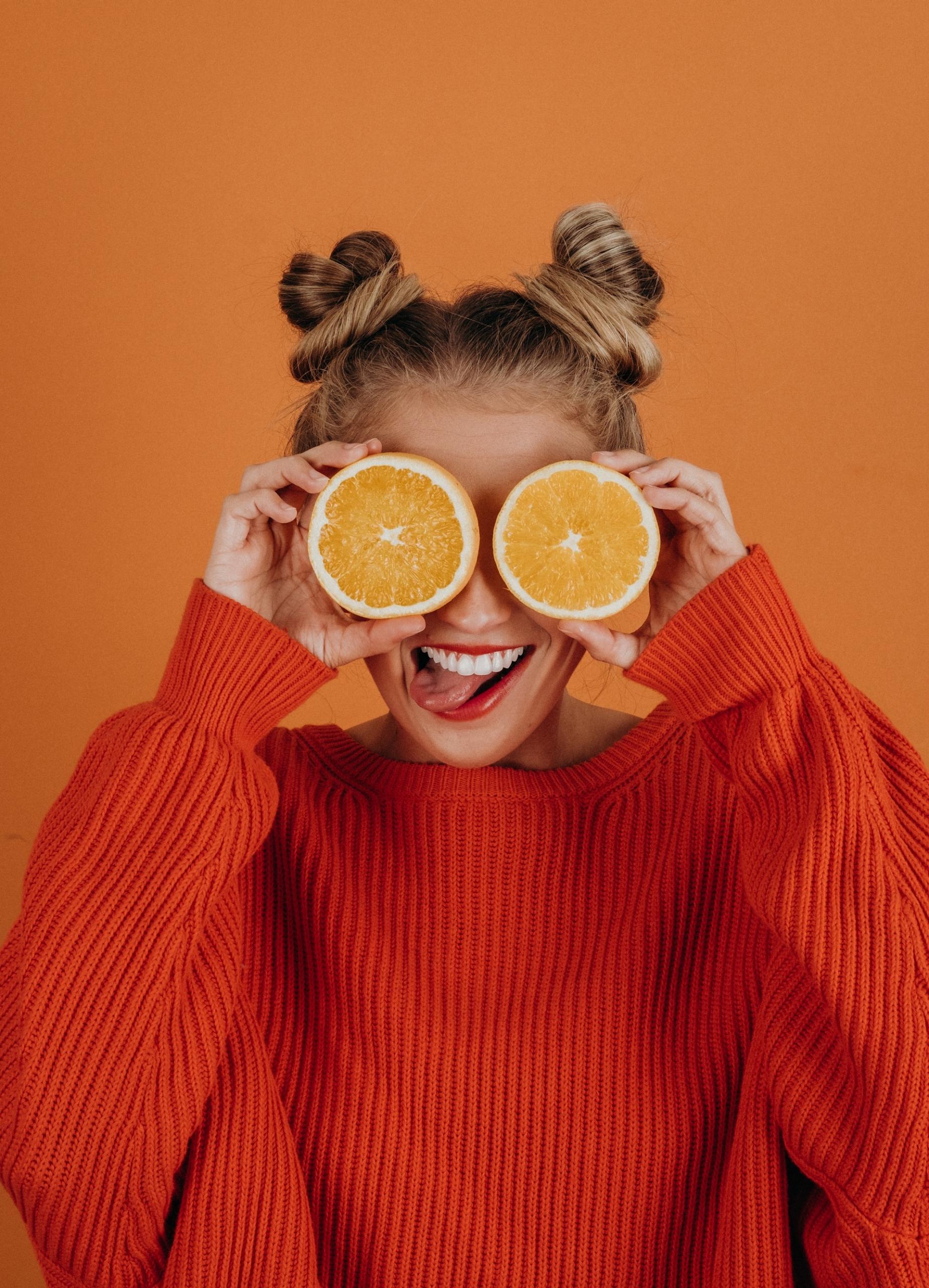 A joyful woman holds halves of an orange over her eyes, embodying the vibrant spirit of a healthy diet that enhances singing performance.