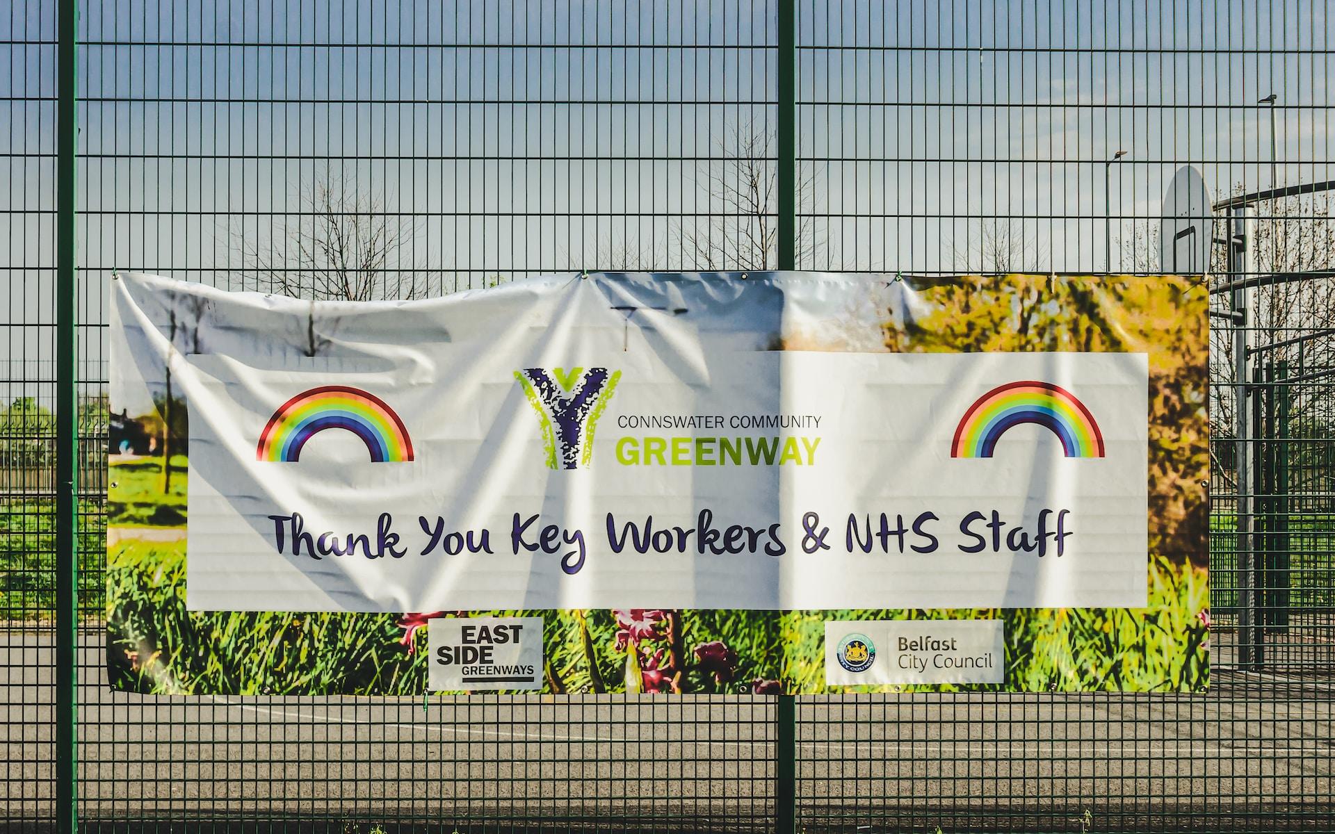 A white banner thanking key workers and NHS in large black letters with rainbows in the upper corners hangs on a fence on a sunny day.