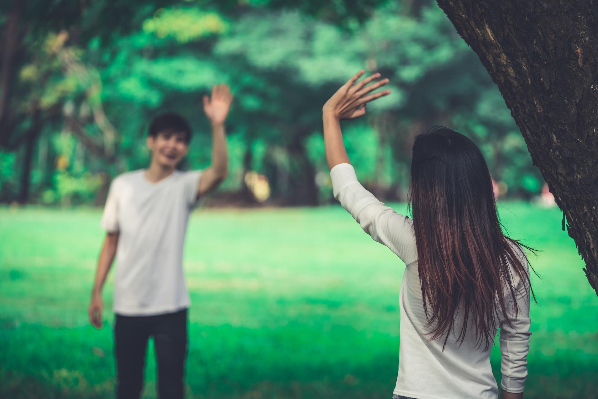 Two people wearing white tops wave at each other from a distance in a green outdoor area, maybe a park.