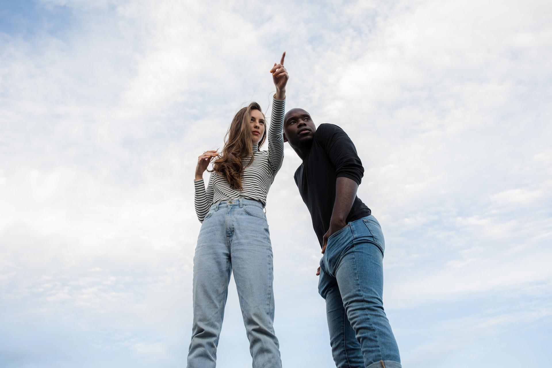 A person wearing light coloured jeans and a long-sleeved striped shirt points as the person next to them, wearing a black shirt and dark jeans, looks in that direction.
