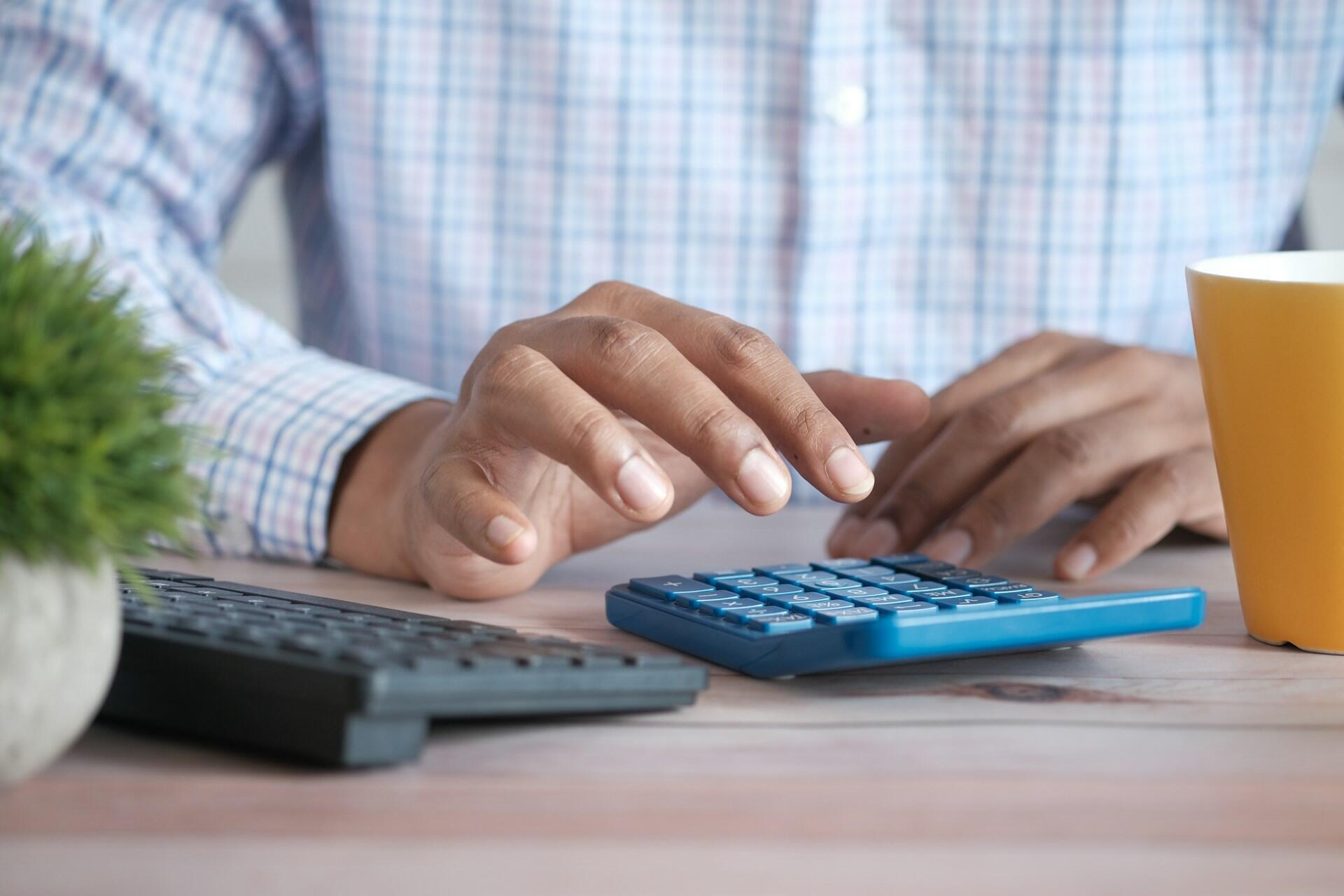 Hands using a calculator on a desk next to a keyboard and a cup