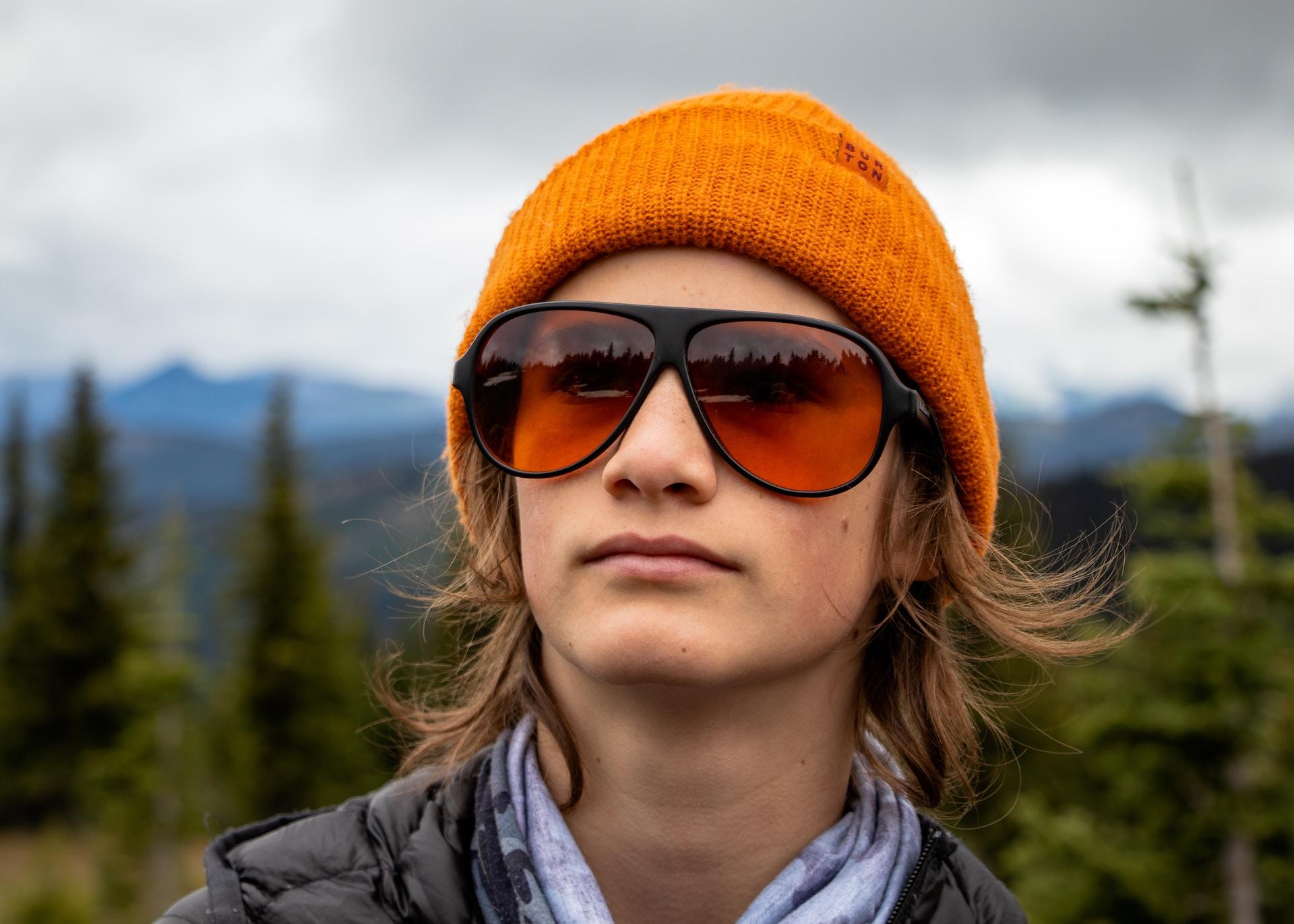 A stylish person wearing an orange beanie and large sunglasses with red lenses stands in a nature area, looking thoughtful.