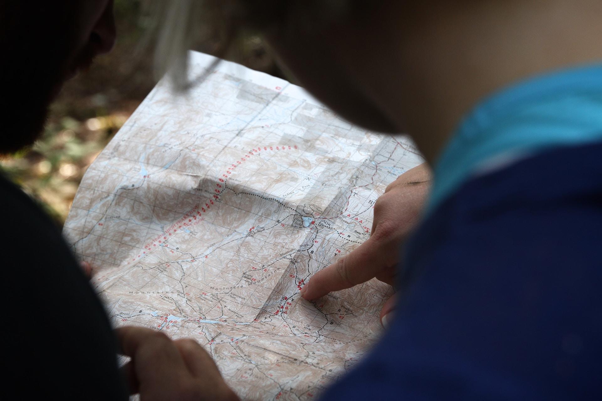 A person wearing a blue top points at something on a mostly brown city map with their right index finger while a person wearing a black top looks on.