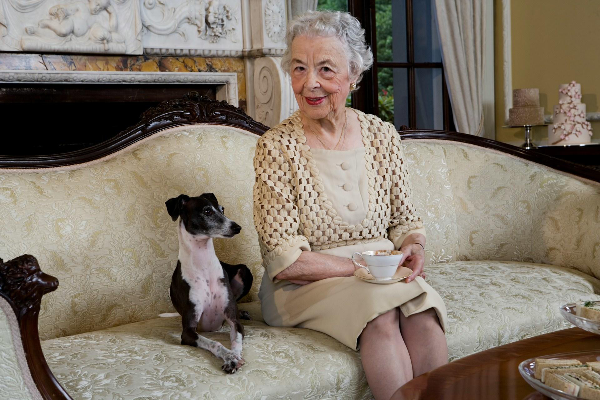 An elderly woman wearing a tan coloured dress with gold accents sits primly on a gold-thread sofa, holding a fine china cup with saucer on her knee, with a black and white dog posed next to her.