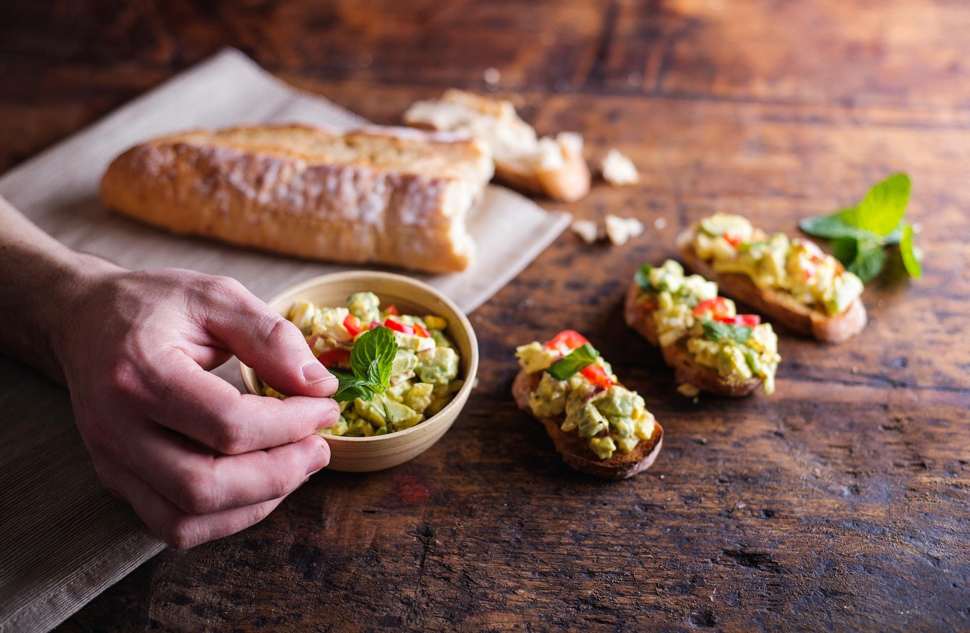 Three slices of bruschetta topped with a green and yellow 'salad', resting on a well-used wooden board, with the rest of the bread on a tan-coloured napkin and a person placing a sprig of green atop a bowl of bread topping.