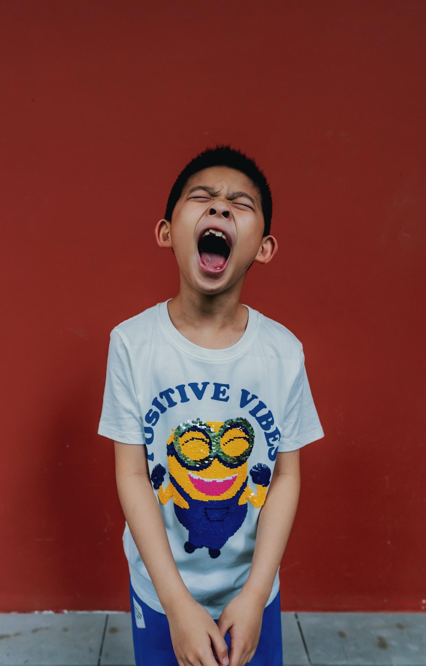 A boy wearing a white tee shirt with a minions design and blue shorts stands in front of a wall painted ochre, screaming.  