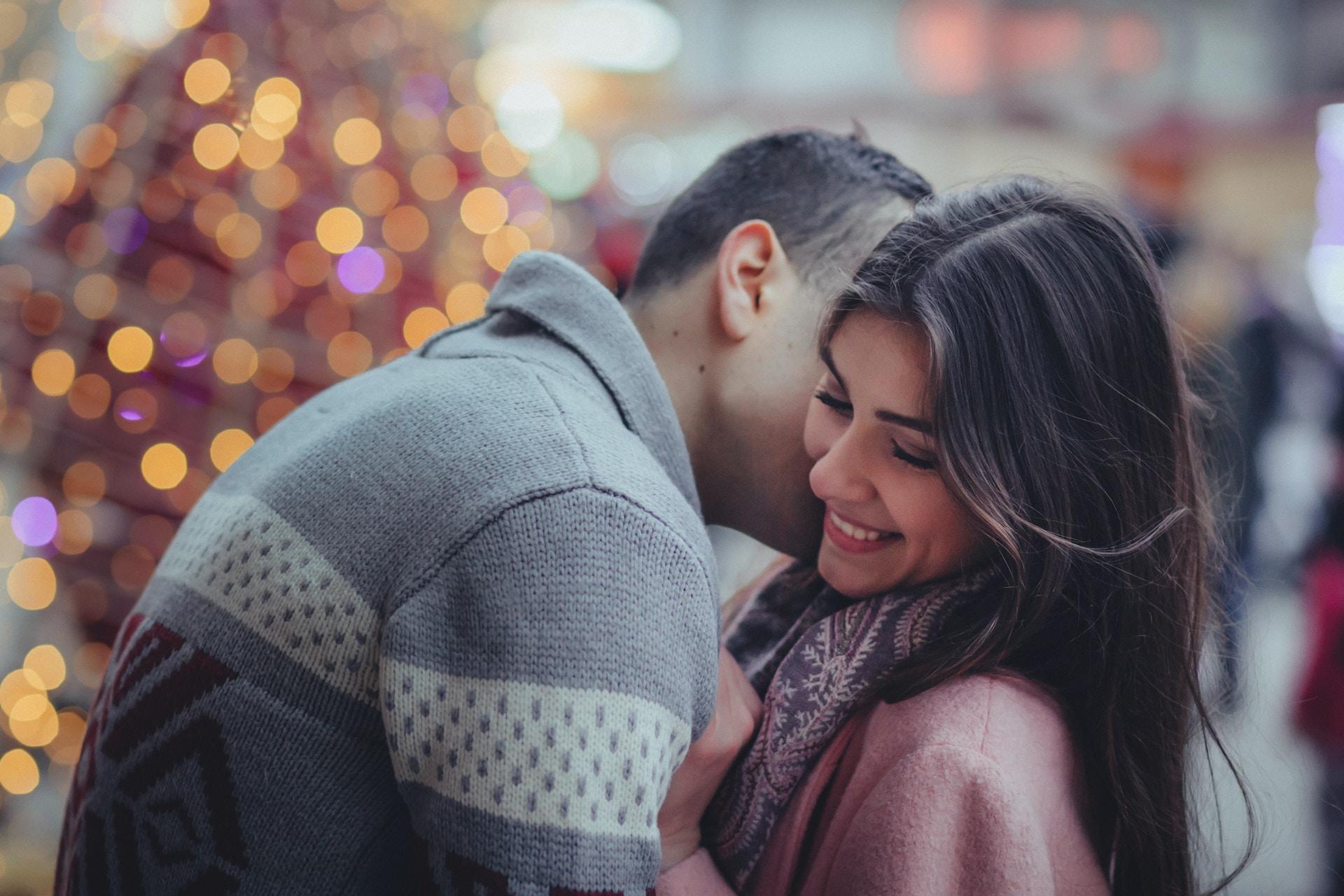 A tall person wearing a grey jumper bends to nuzzle a person wearing a pink jumper with a brown scarf in front of a blurred-out, lit-up decoration.