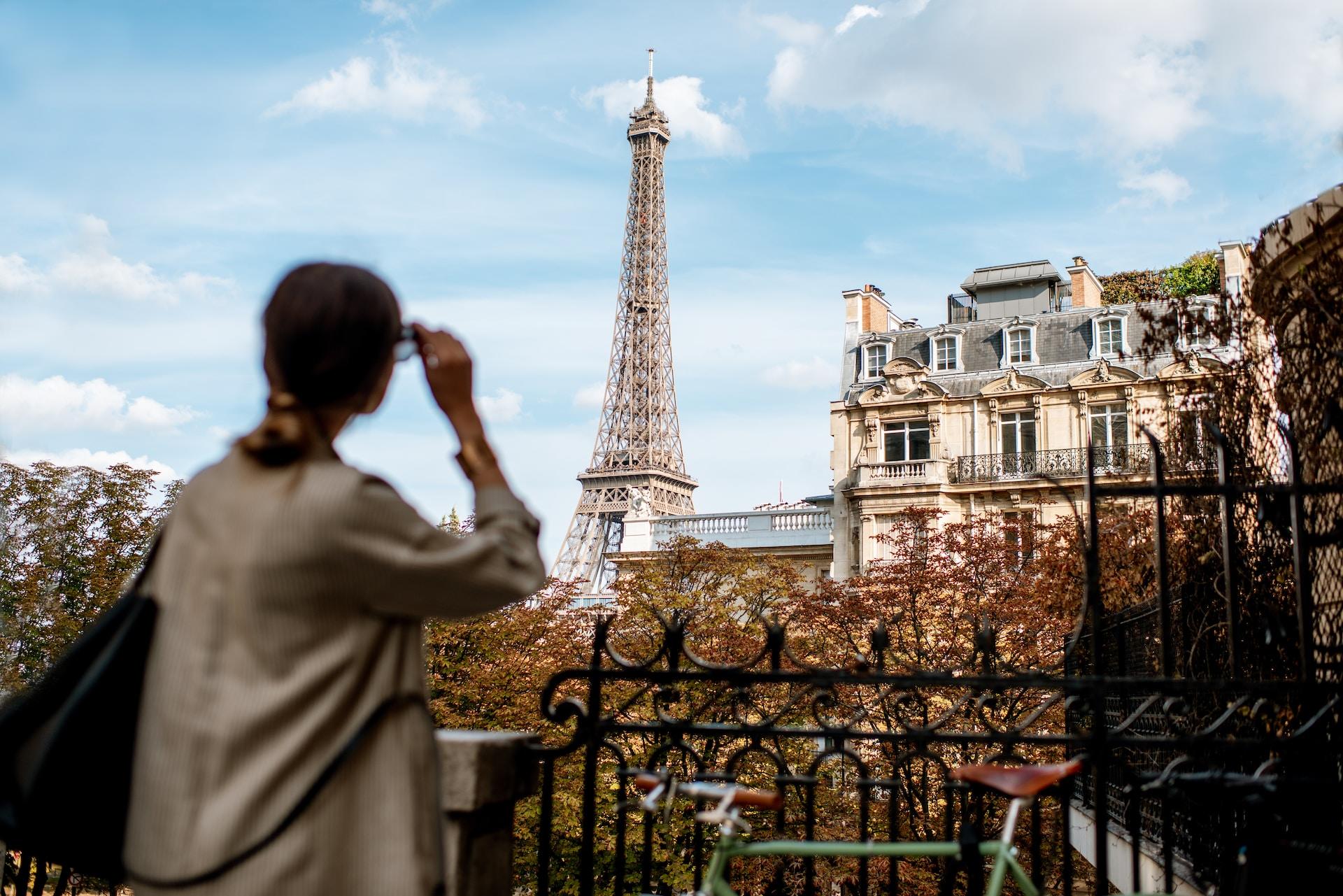 A person wearing a light coloured loose top raises their eyeglasses to look at the Eiffel Tower in the distance.