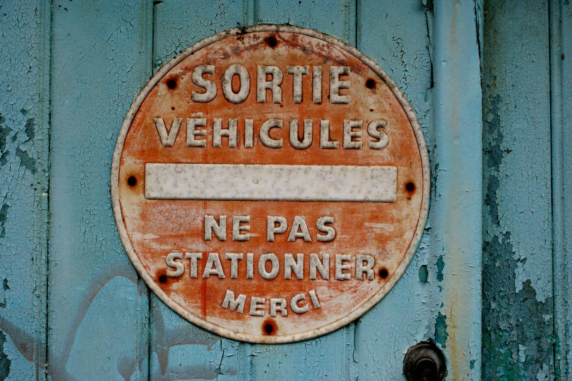 A faded red sign on a blue door marked with chipping paint that warns of exiting cars and urges no parking, thank you.