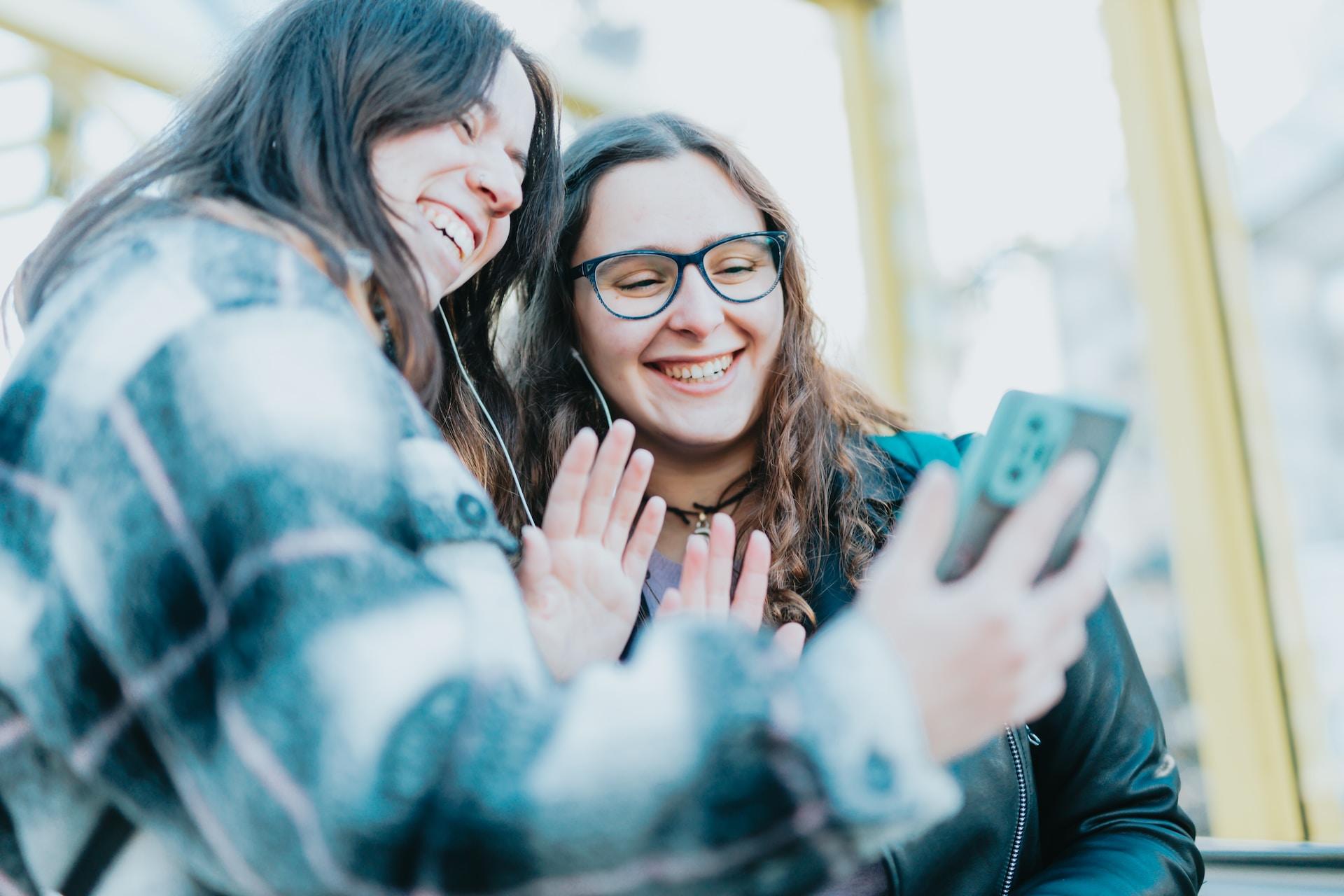 Two people wearing cold-weather clothing smile and wave at a smartphone's screen.