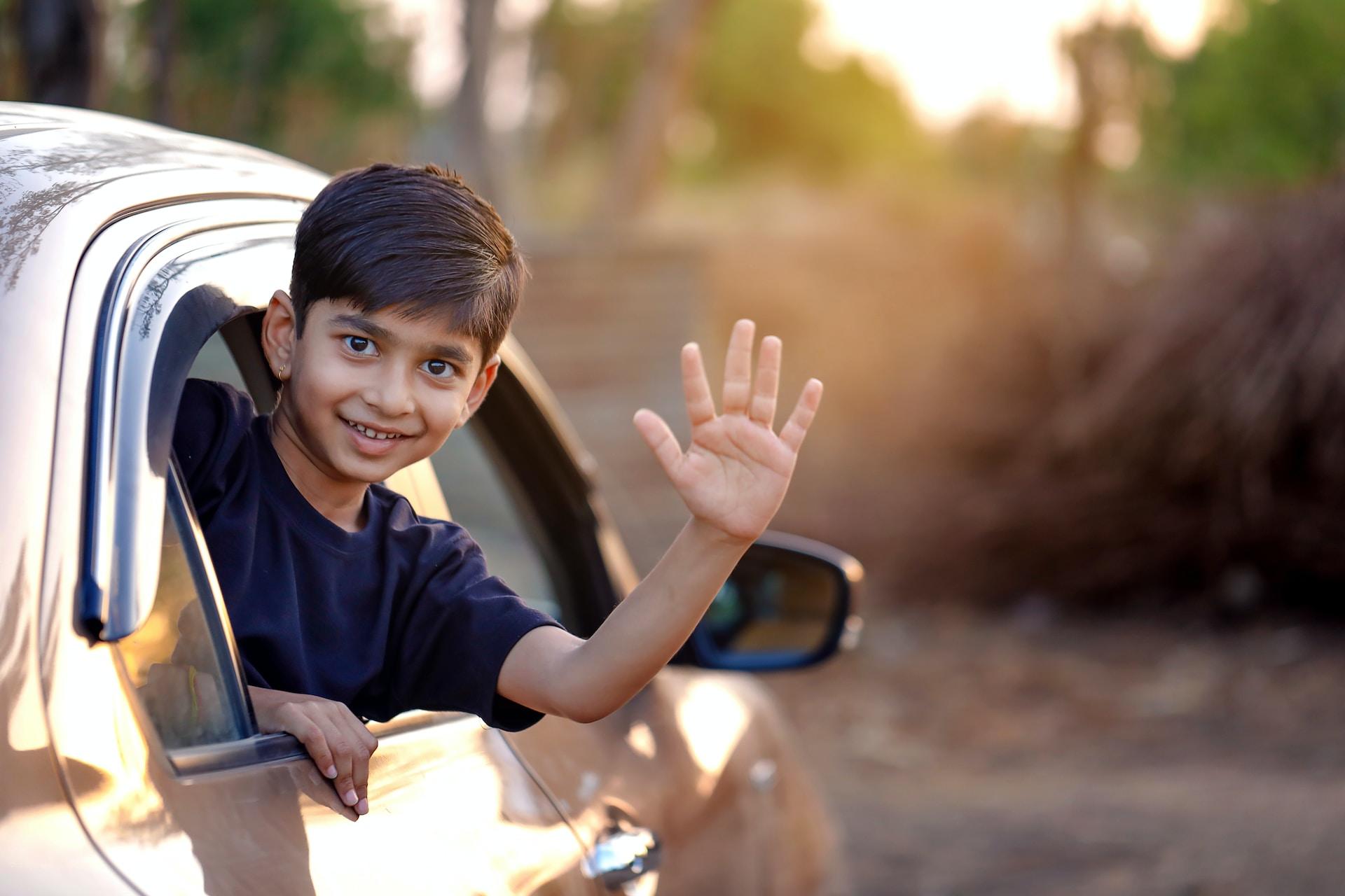 A boy wearing a black tee shirt leans out of a car's rear passenger window and smiles as he waves goodbye while the sun bathes the scene in golden light.