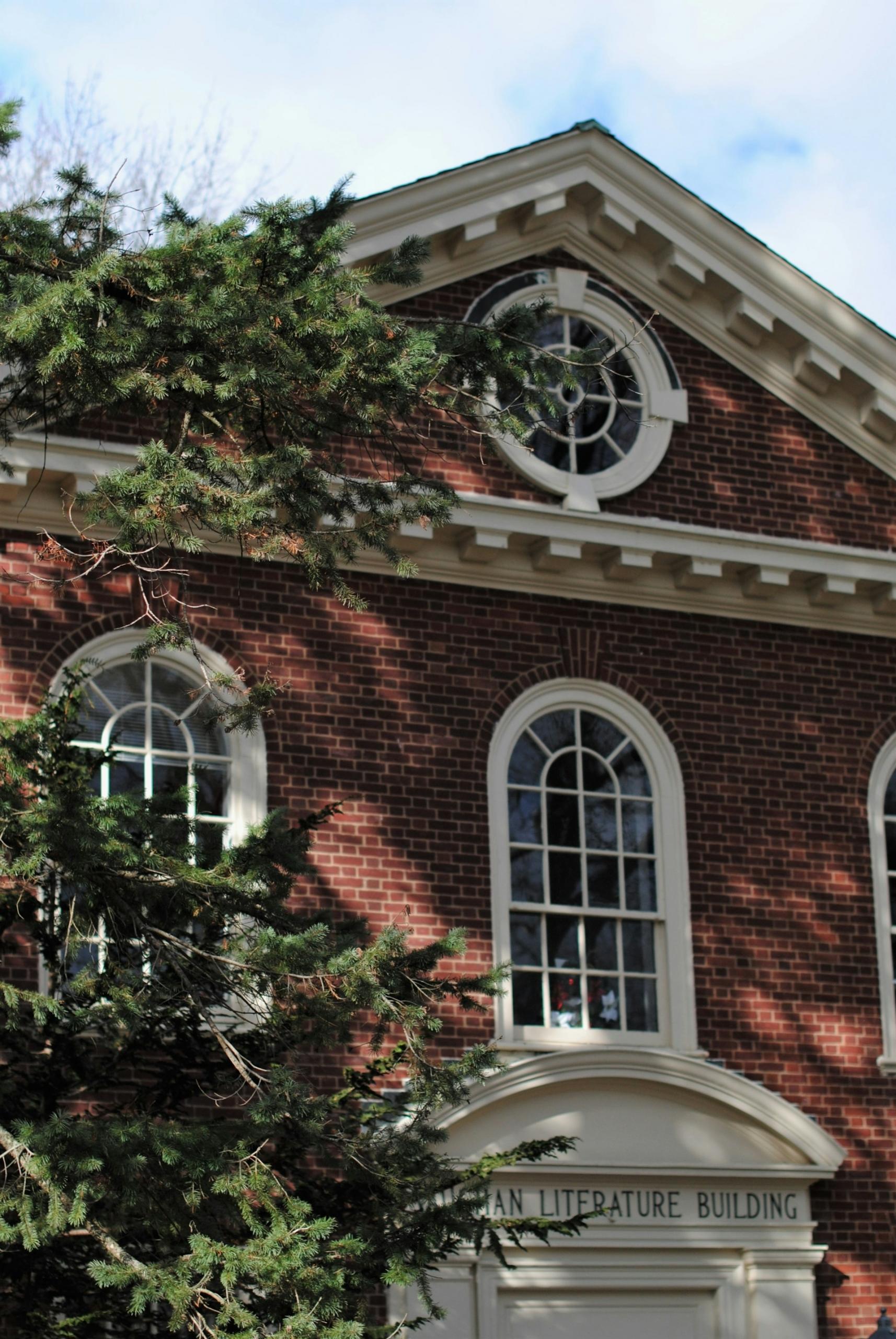 A building with a red brick facade and ornate windows and entranceway framed in white stands in contrast to the green tree which casts its shadow on a sunny day.