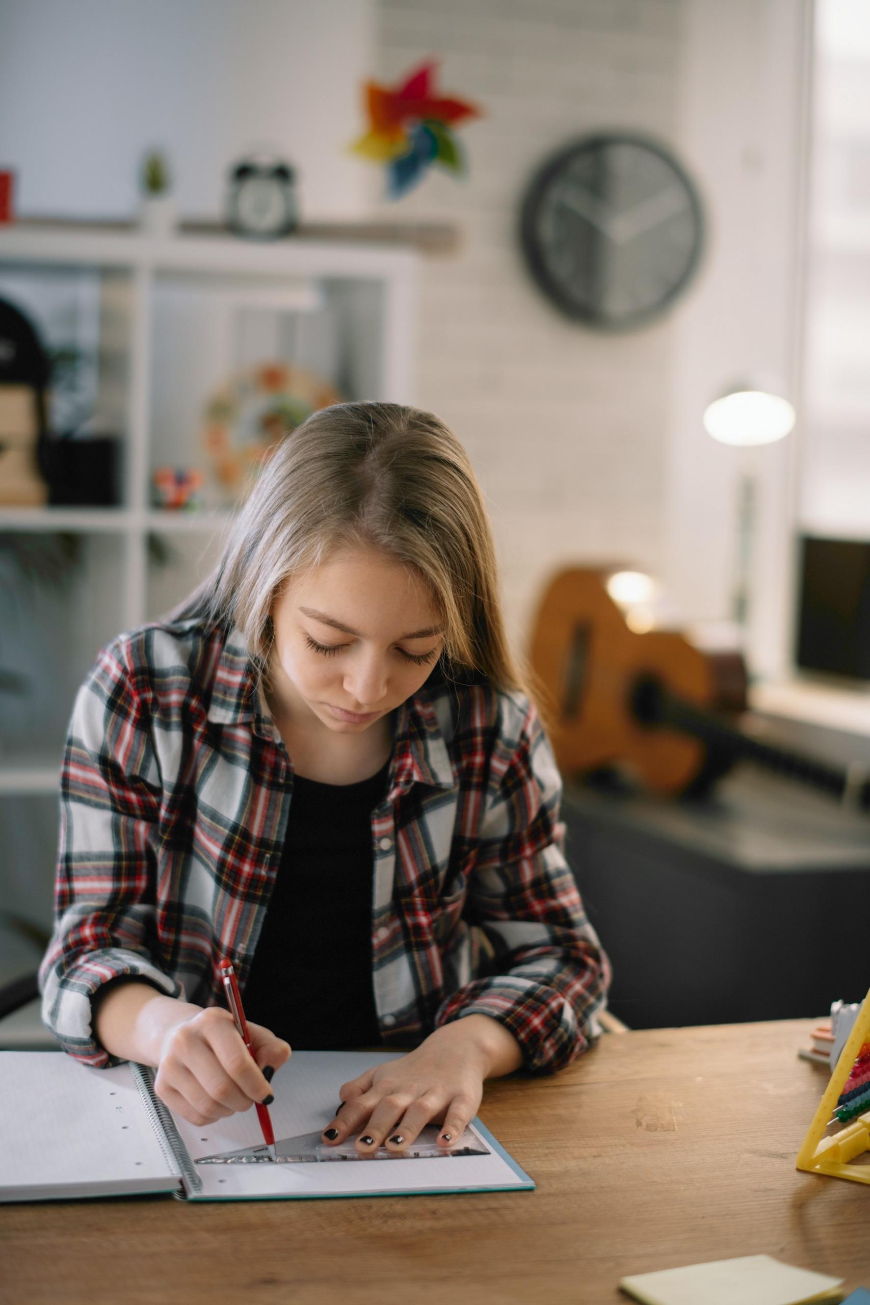 A child wearing a plaid red, white and black top over a black tee sits at a wooden table in a bright room, holding a square on an open notebook page with their left hand while their right traces a line with a red pen.