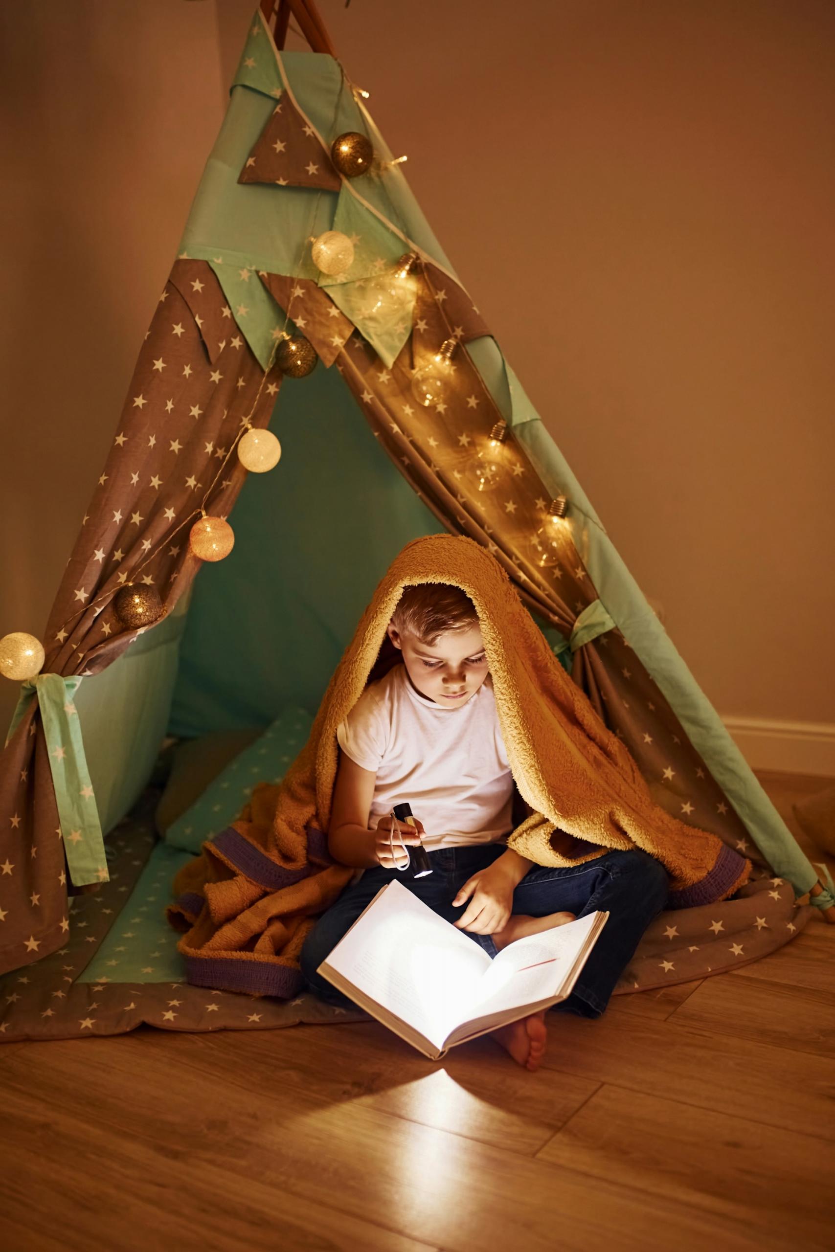 A child a white tee shirt and dark trousers sits in a green and brown play tent with a string of large round lights draped around the entrance. They have a blanket on their head and shine a lit torch on the pages of an open book.