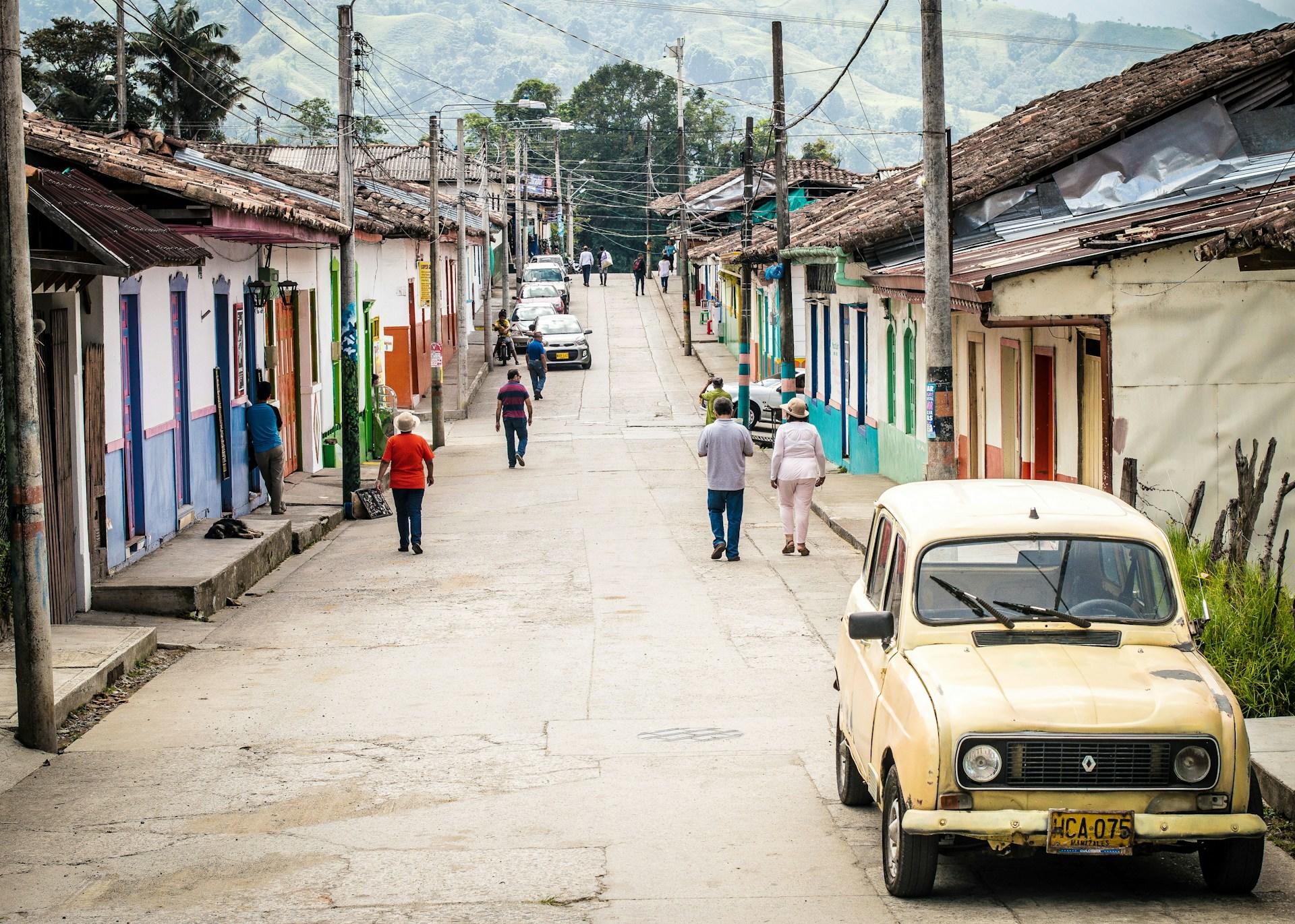 A Colombian town, people are walking down the street, there is a yellow vintage Renault in the forefront of the image.